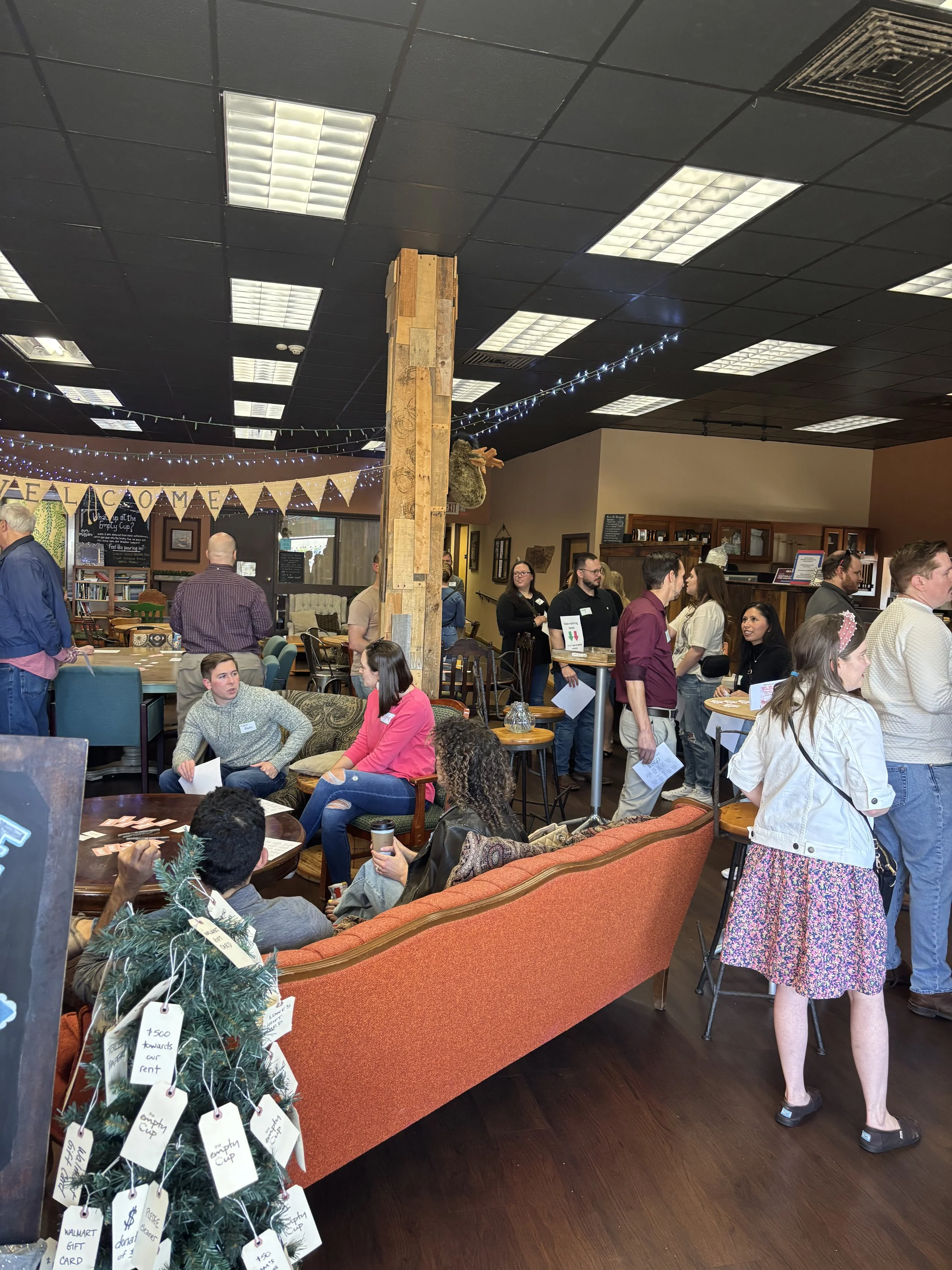 People gather in a cozy, decorated cafe or community space. Some people sit on chairs and talk, others stand in line or socialize. There is holiday decor, including a small Christmas tree with donation tags and a hanging reindeer, and string lights. The room has a dark ceiling with fluorescent lighting and wood flooring.