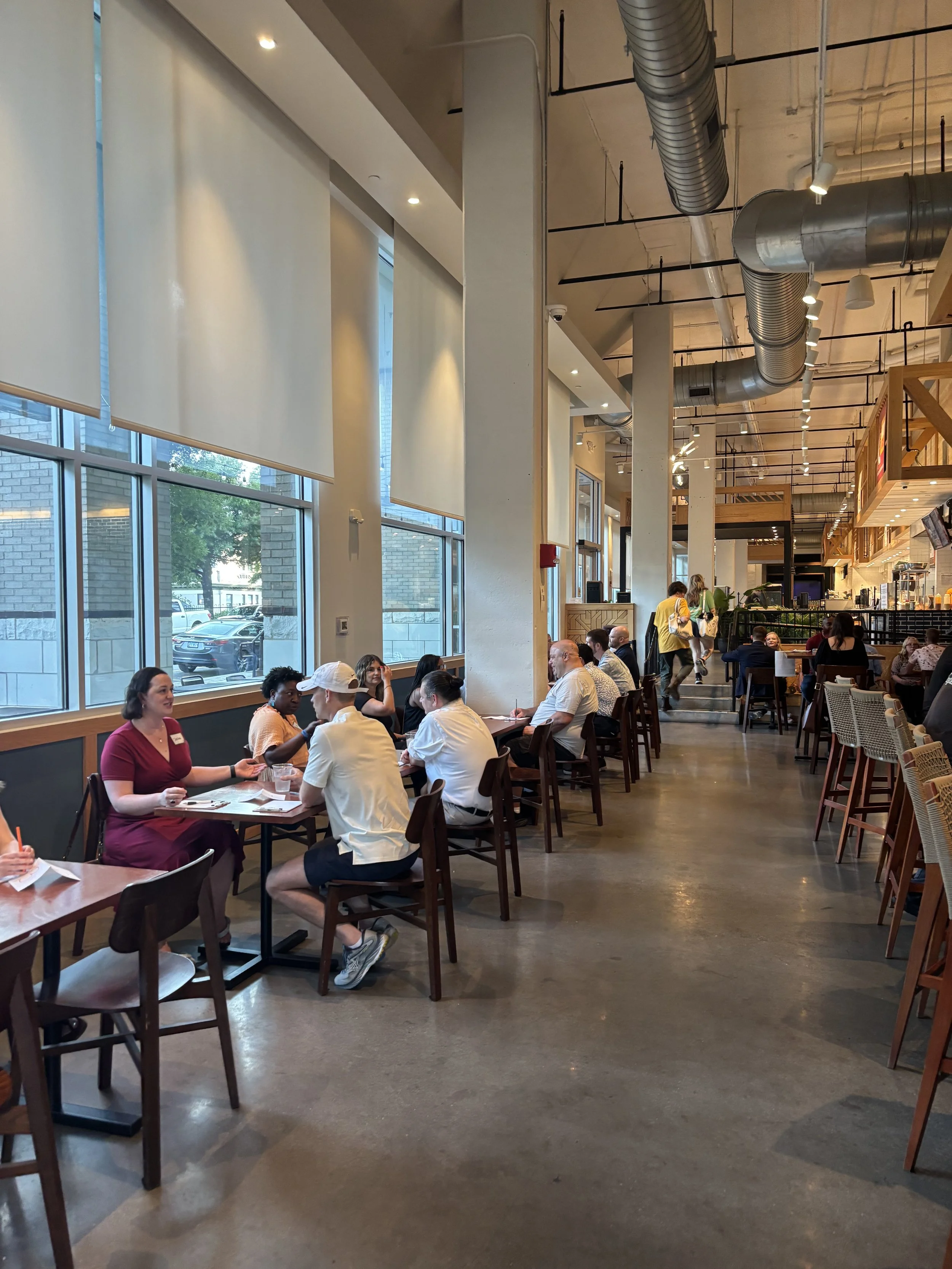 People sitting at tables and talking inside a modern restaurant or cafe with large windows and industrial ceiling pipes.