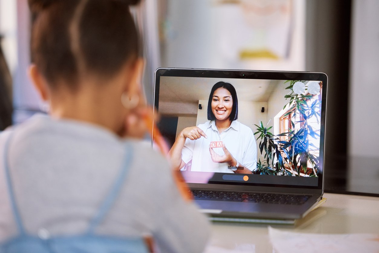 A girl looks at a laptop screen showing a virtual speech therapist smiling and holding up colorful blocks during an online speech therapy for children in Michigan.