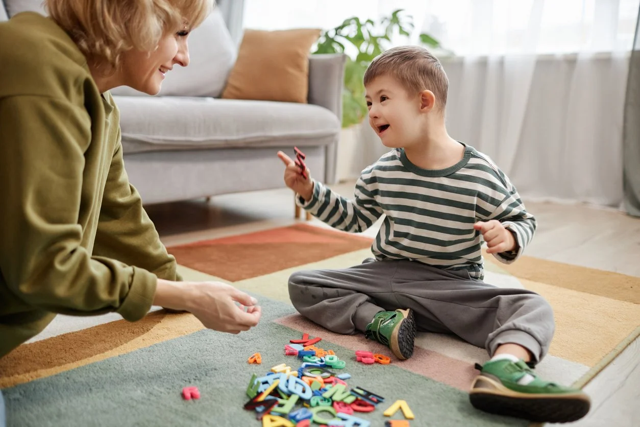 A young boy and a speech therapist sitting on a colorful rug, playing with foam alphabet letters indoors while working on language skills during speech therapy for children in Whitmore Lake, MI.