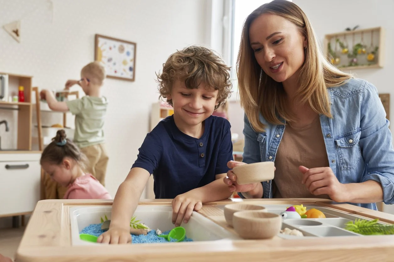 A school contract occupational therapist and a boy playing with a sensory table filled with blue sand and various toys in a classroom during occupational therapy for kids in Michigan.