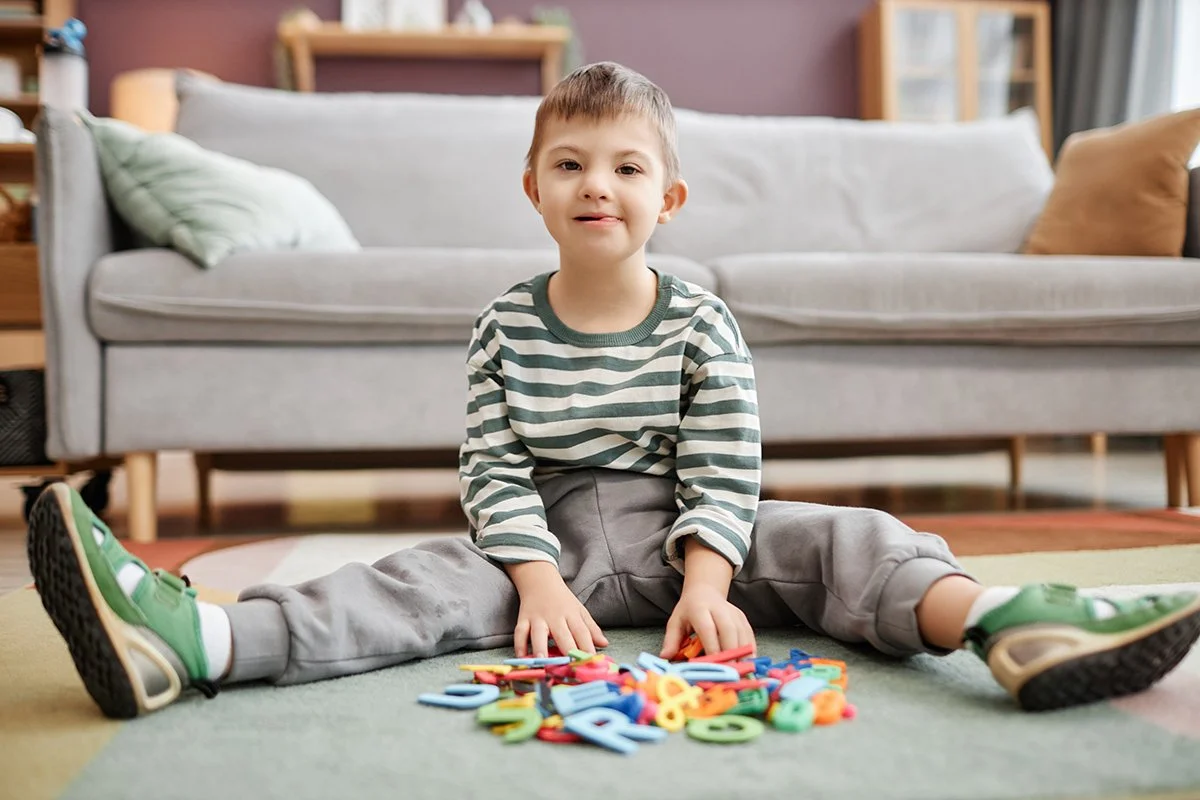 A young boy with developmental delays is sitting on the floor in front of a couch with colorful foam letters and numbers, smiling at the camera during speech therapy for children in Fenton, MI.