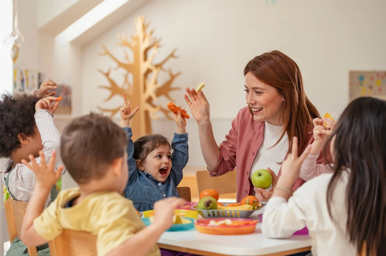 A school contract speech therapist is smiling and holding an apple interacts with children sitting around a table in a brightly lit room decorated with a tree-shaped wall ornament.