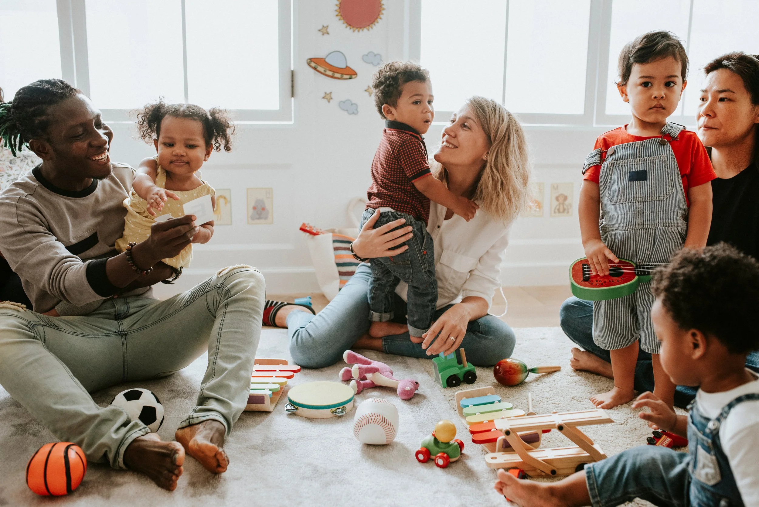 A group of diverse children and their parents interacting on a carpeted floor and playing with toys, music instruments, and sports balls during a play group for young children to work on early developmental skills in Fenton, MI.