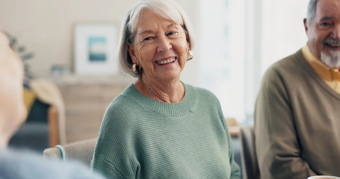An elderly woman with gray hair smiling and wearing a green sweater, sitting with others and communicating with improved loudness after voice therapy for adults in Fenton, MI.
