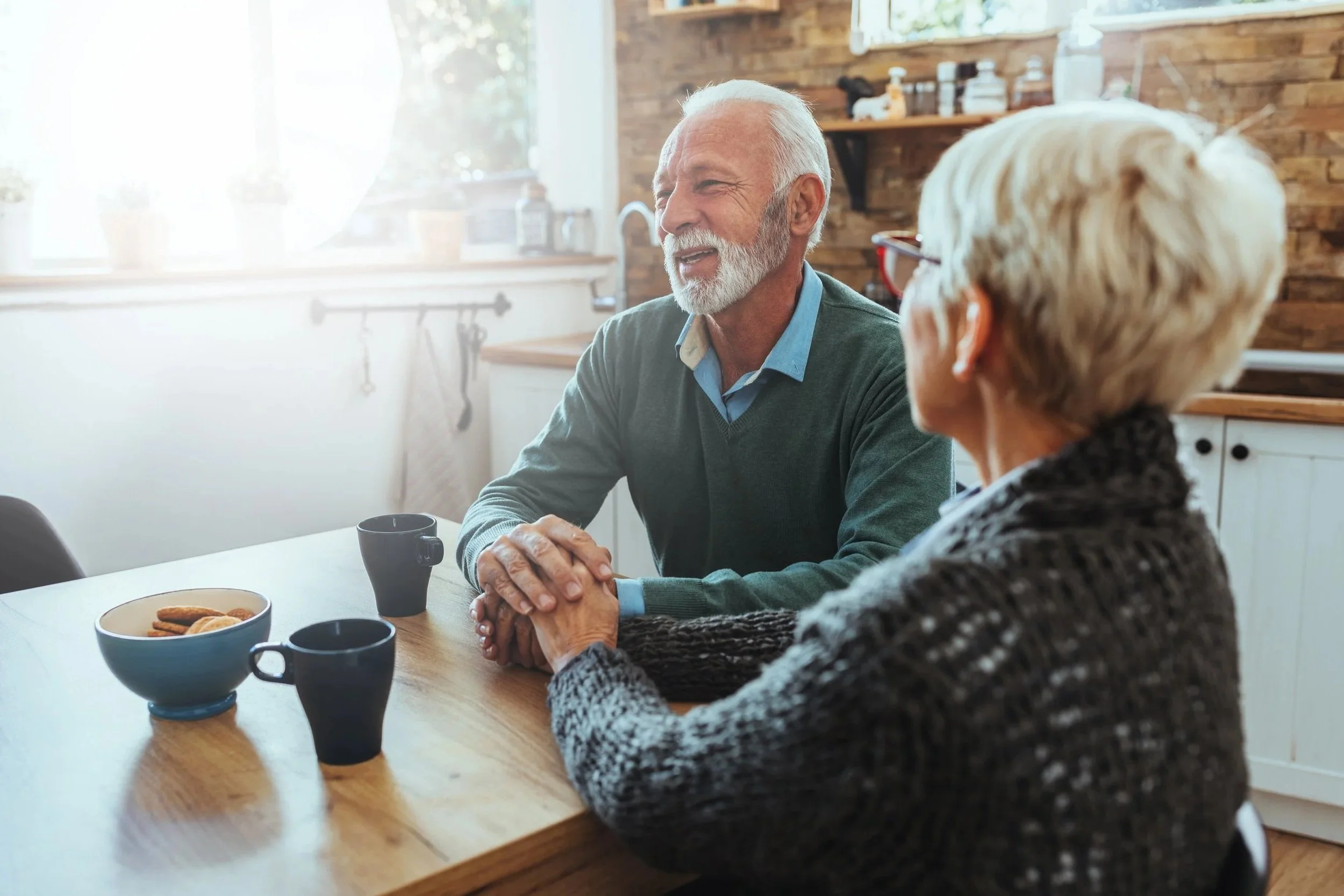 An elderly man and woman sitting at a wooden dining table, holding hands, with coffee mugs in front of them, both with improved swallowing and communication after speech therapy for adults in Fenton, MI.