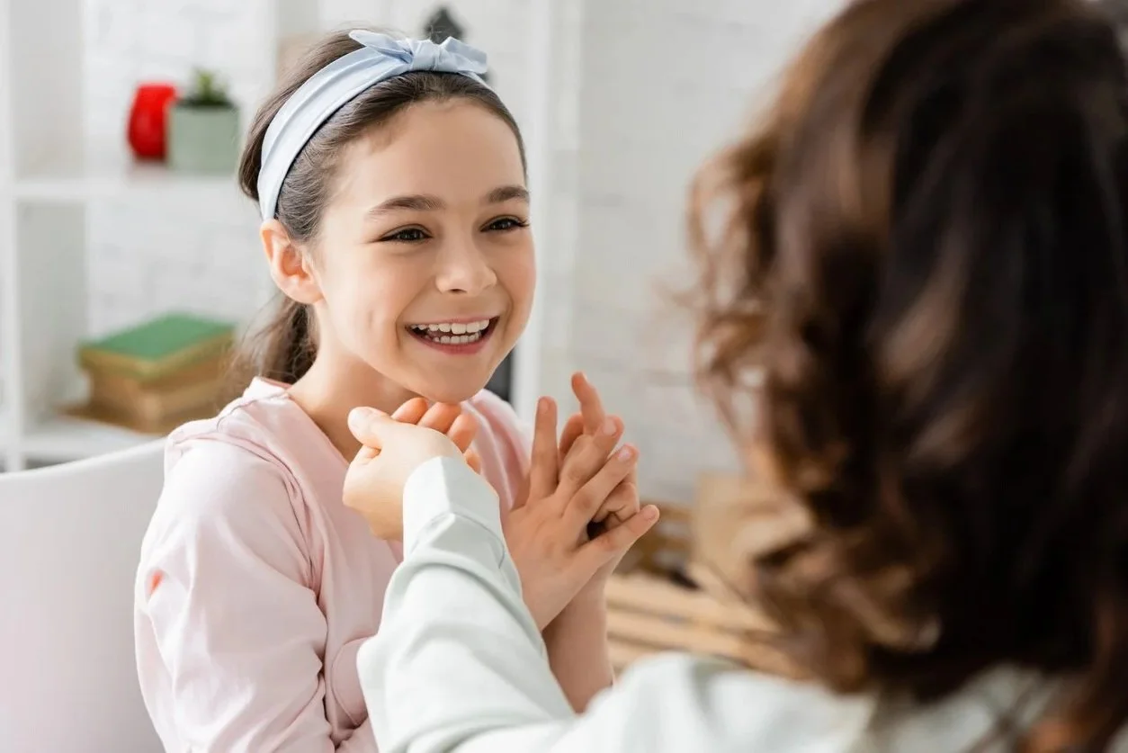 A young girl with a gray headband smiling and engaging in a conversation with her speech therapist while working on speech sounds during speech therapy for children in Fenton, MI.