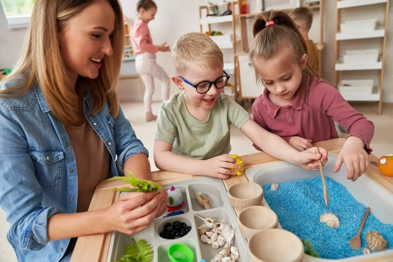 A school contract occupational therapist in Michigan is playing with two children at a sensory table filled with blue rice, seashells, and small toys.