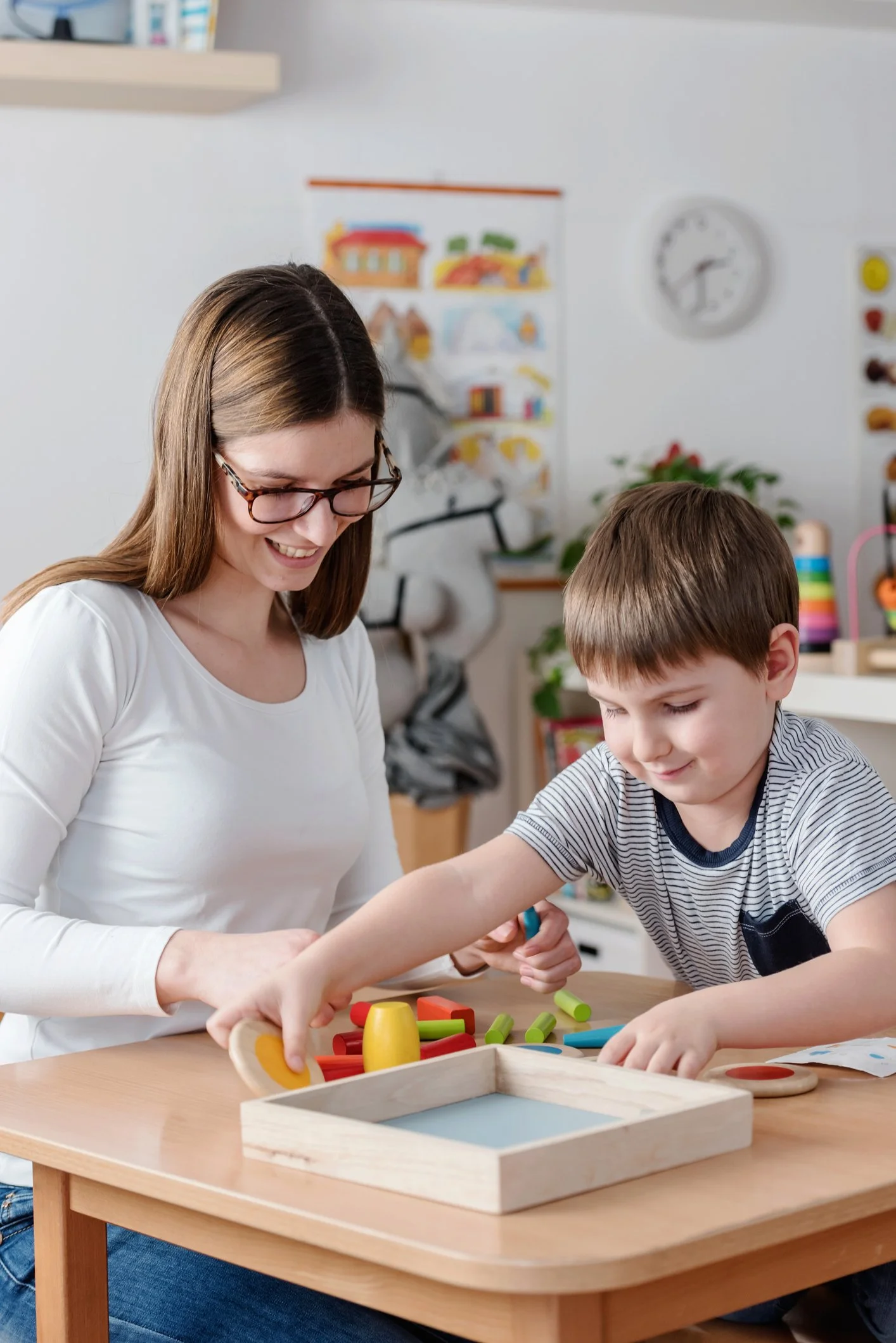 A speech therapist and a young boy are playing with colorful wooden toys at a table in a playroom. The boy is receiving speech therapy for language skills in Fenton, MI.