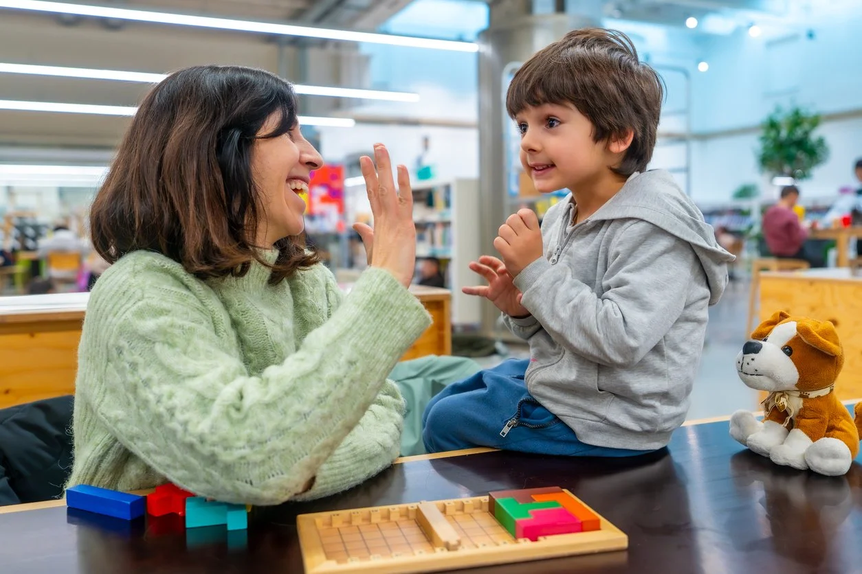 A speech therapist and a young boy playing a game at a table during speech therapy for kids in Whitmore Lake, MI. A stuffed dog toy is on the table, and bookshelves can be seen in the background.
