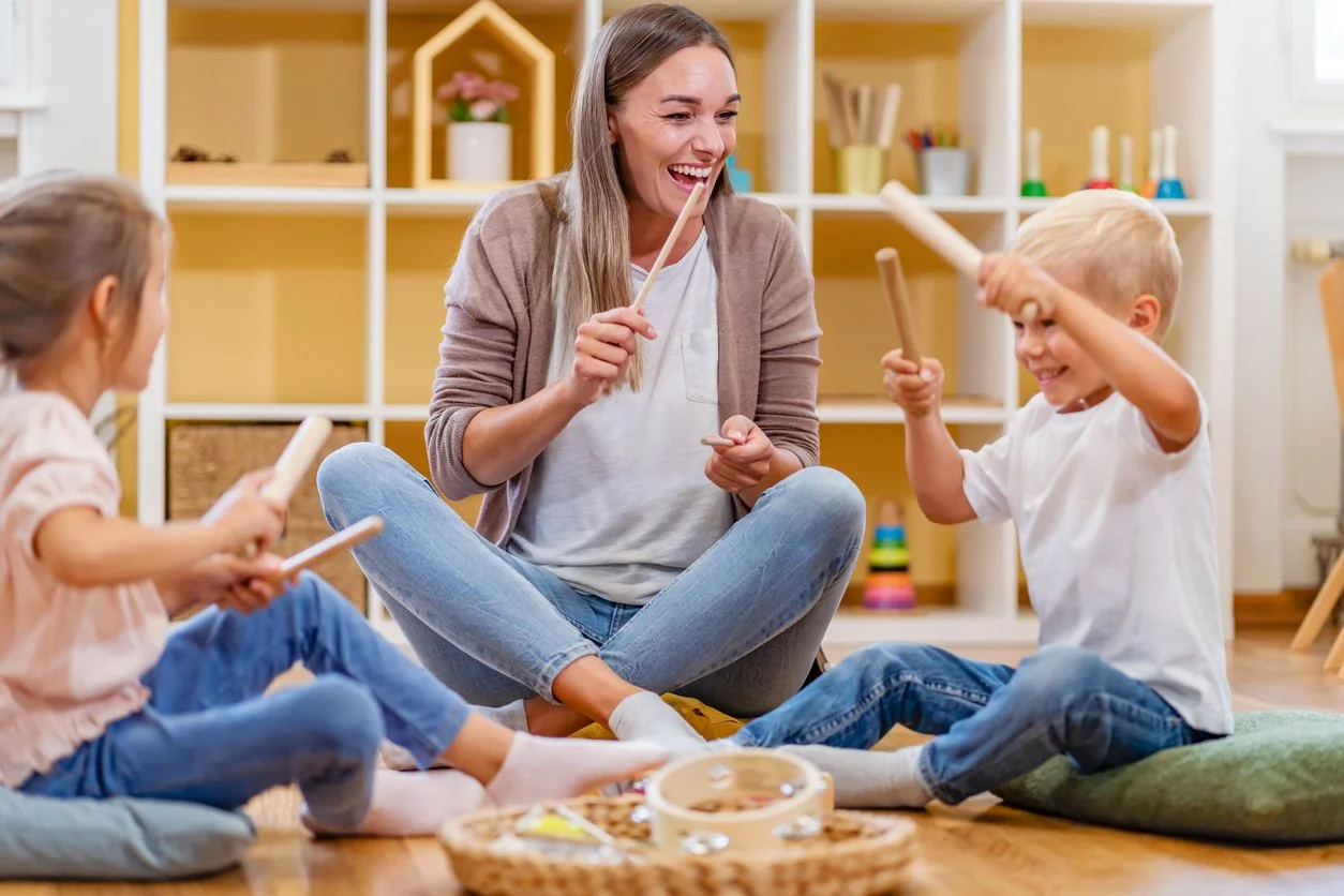 An occupational therapist and two children playing a drumstick game indoors, sitting on the floor with toys and cushions around. The occupational therapist is provided by a therapy contract company in Michigan.