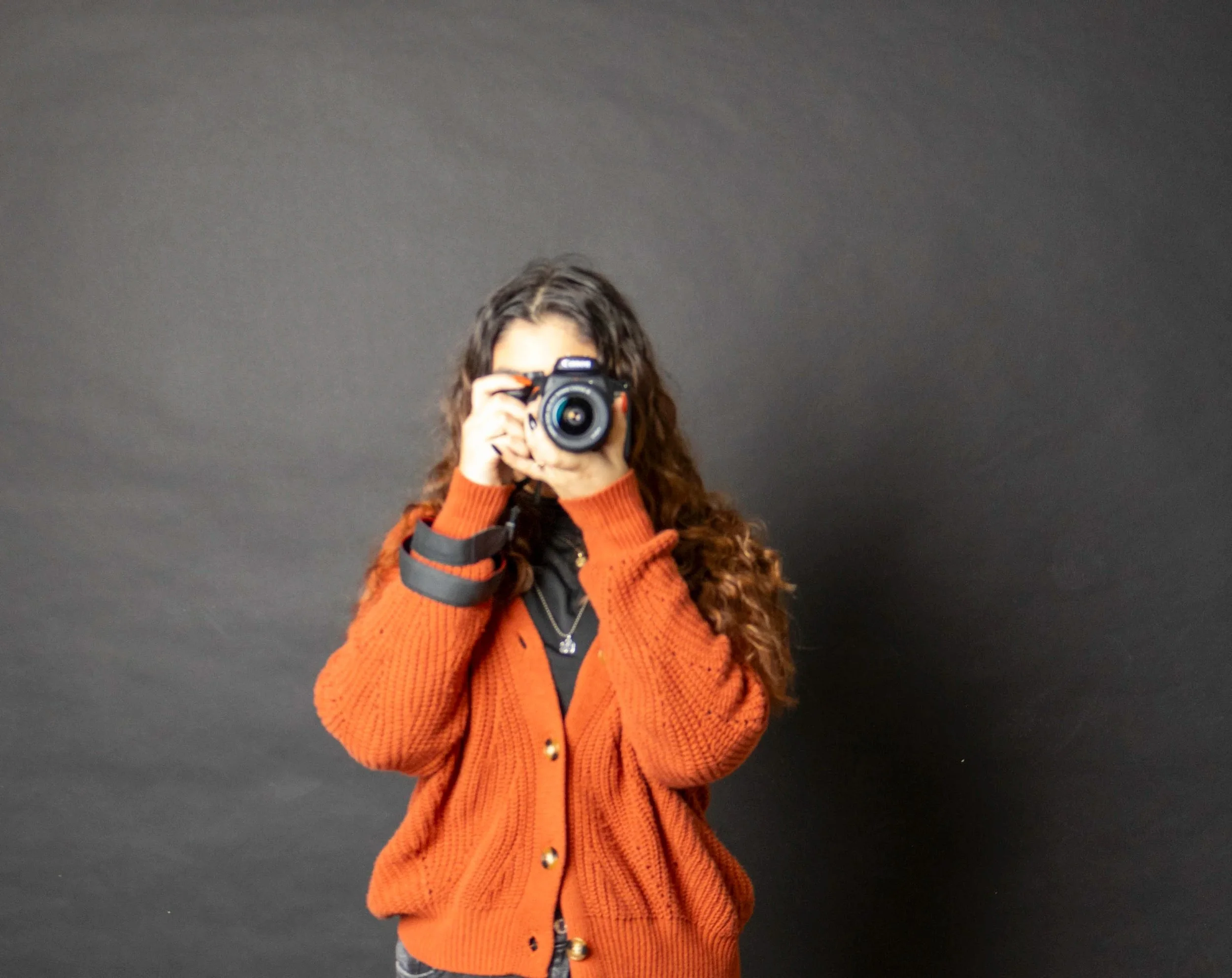 Person with curly hair taking a photo with a camera against a black background, wearing an orange cardigan.