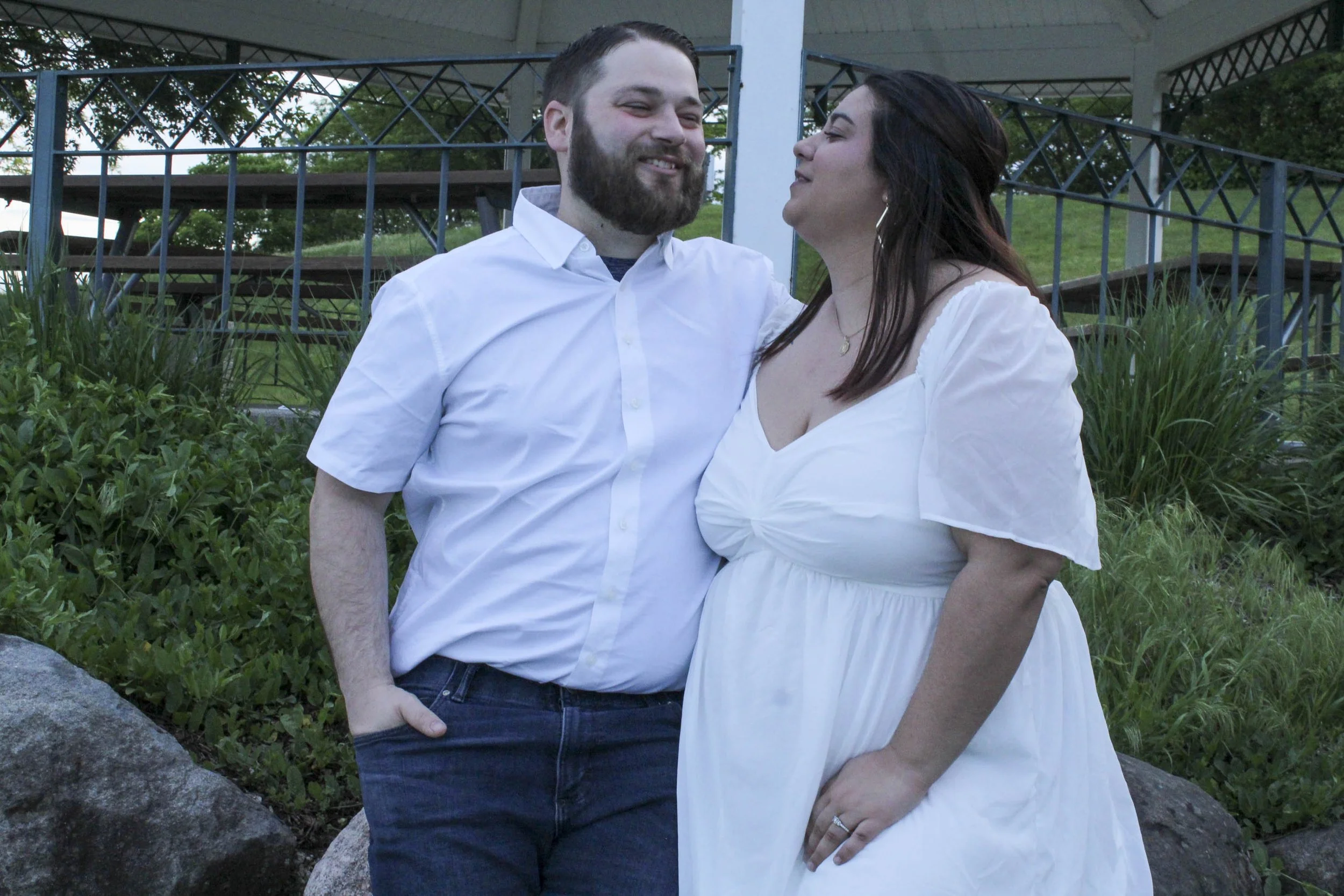 A smiling man and a woman in a white dress share a tender moment outdoors near rocks and greenery, with a pavilion and trees in the background.