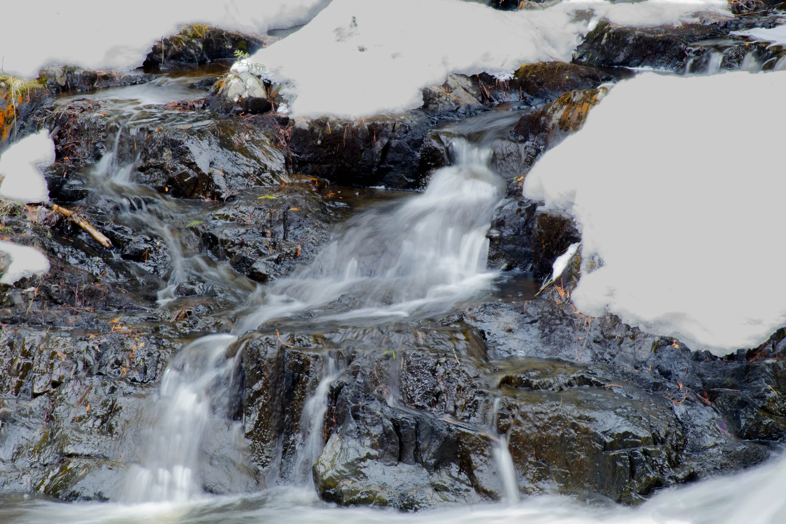 A flowing mountain stream with water cascading over rocks, some moss-covered, with patches of snow on the rocks around it.