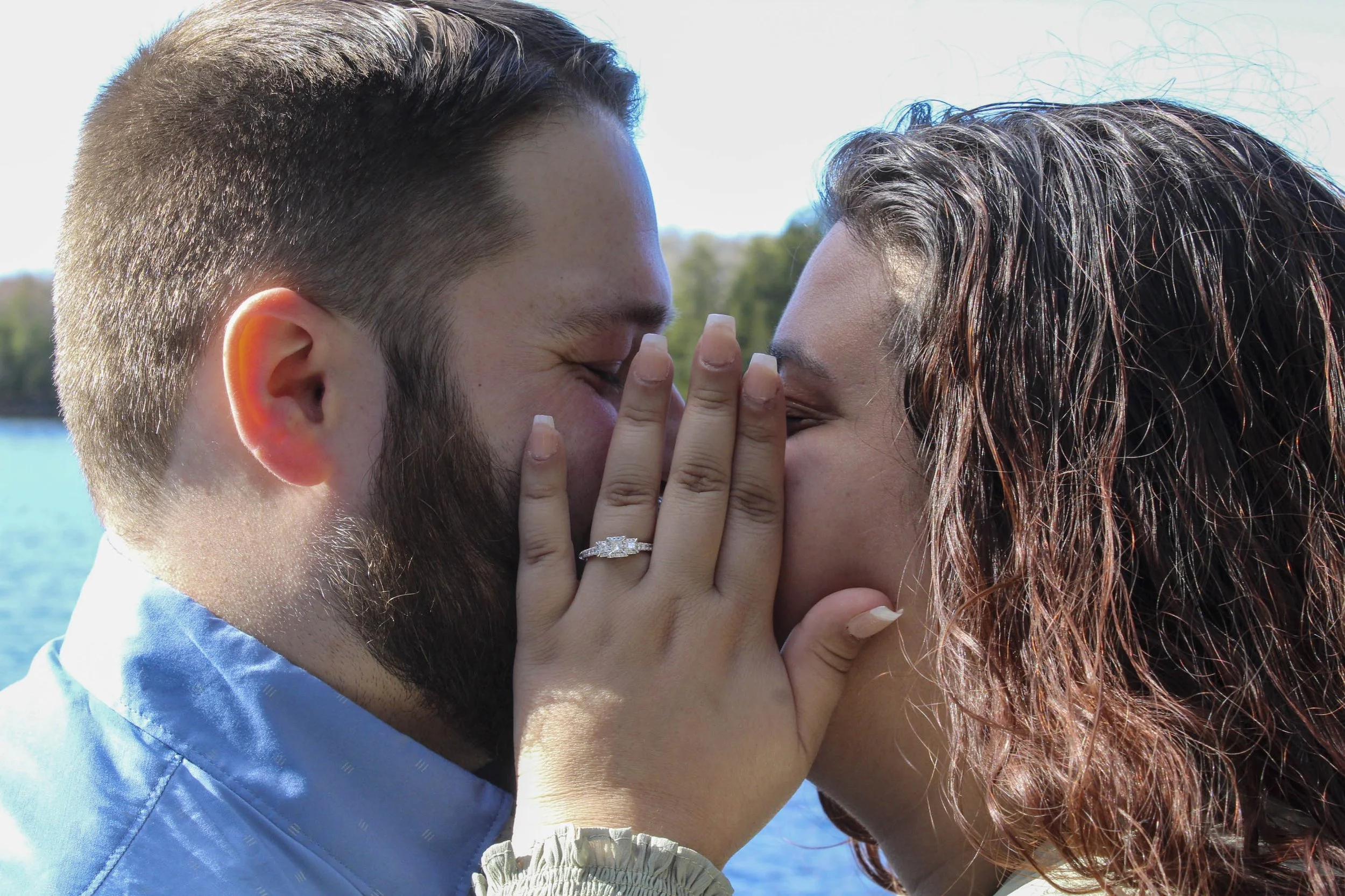 A man and woman share an intimate moment, with their faces close together, outside near a body of water, with the woman touching the man's face and wearing an engagement ring.
