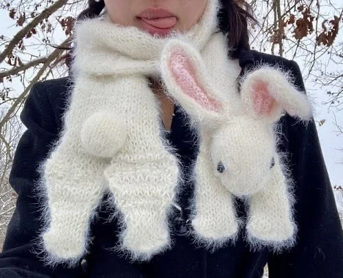 Person wearing a black coat with two white knitted scarves shaped like animal figures, one resembling a bunny with pink ears and the other a small bear, hanging around their necks.