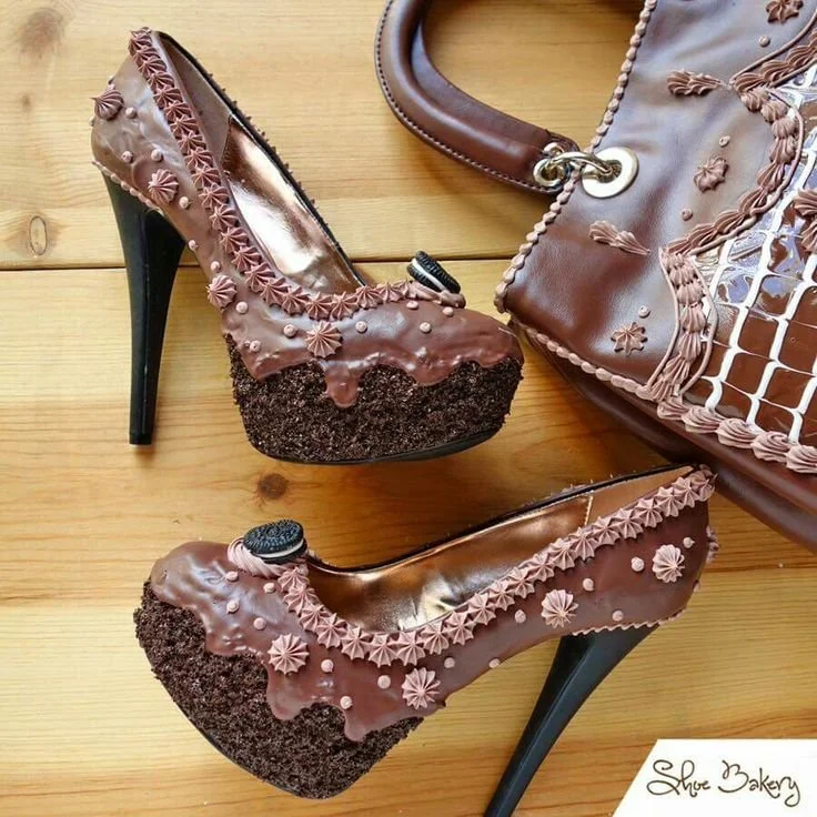 High-heeled shoes designed to look like chocolate cake with icing and sprinkles, placed on a wooden surface, with a matching handbag nearby.