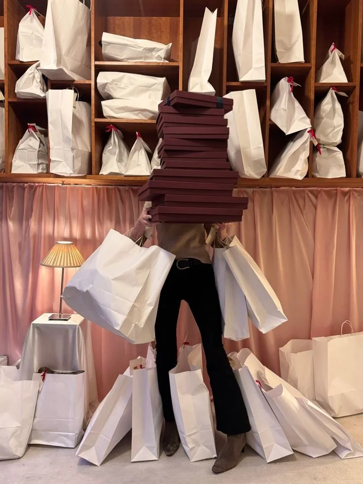 Person standing in front of pink curtains, surrounded by white shopping bags, holding a large stack of boxes above their head, with more bags on the floor and shelves behind.