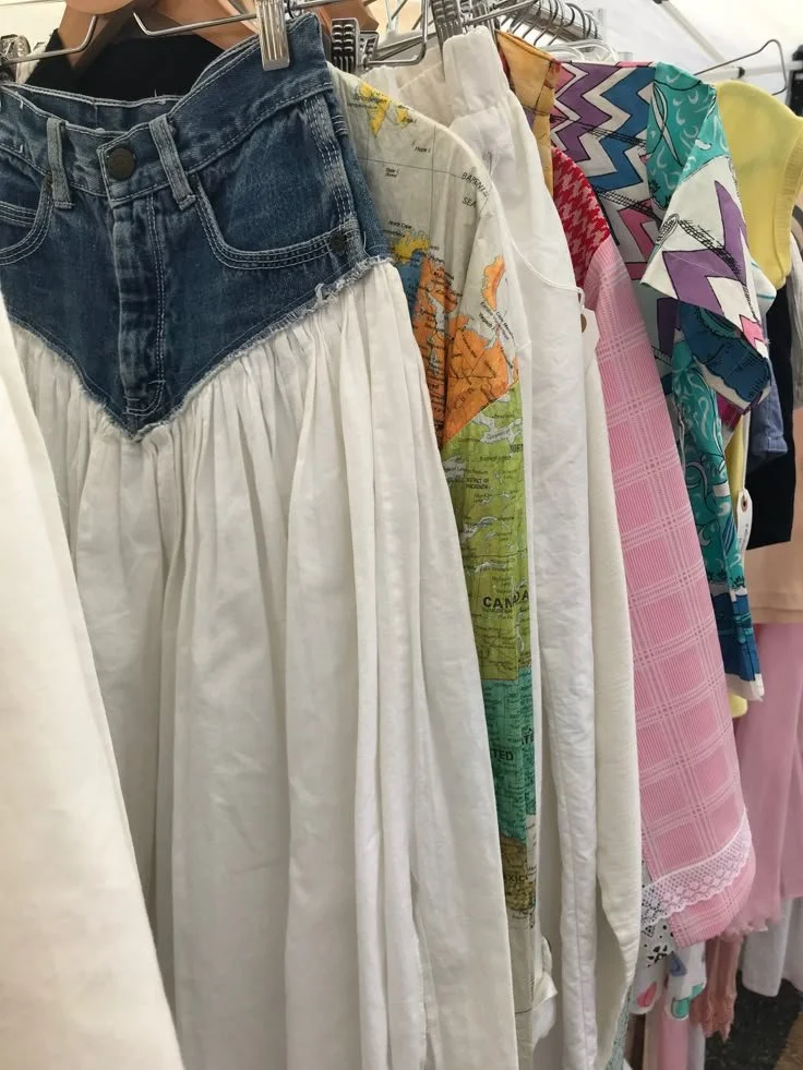 Clothing rack with various garments including a pair of denim shorts with a white skirt attached, a printed shirt, a pink checked dress, and colorful patterned fabrics.