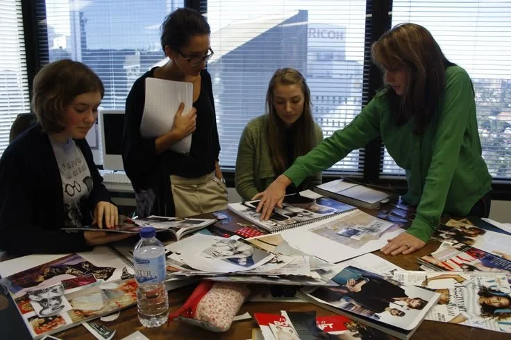 Four women working together at a cluttered office table with magazines, papers, and a water bottle, in front of a window with blinds.