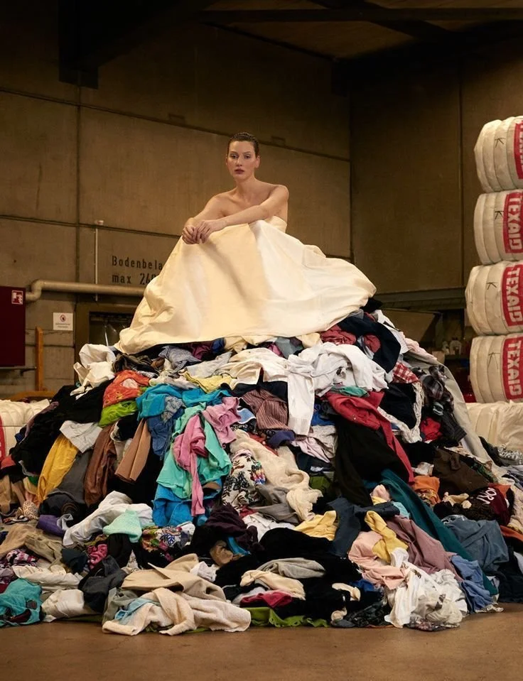 A woman sits on top of a large pile of mixed clothes in an industrial setting. The woman is topless, with her arms resting on the pile, and is looking directly at the camera.