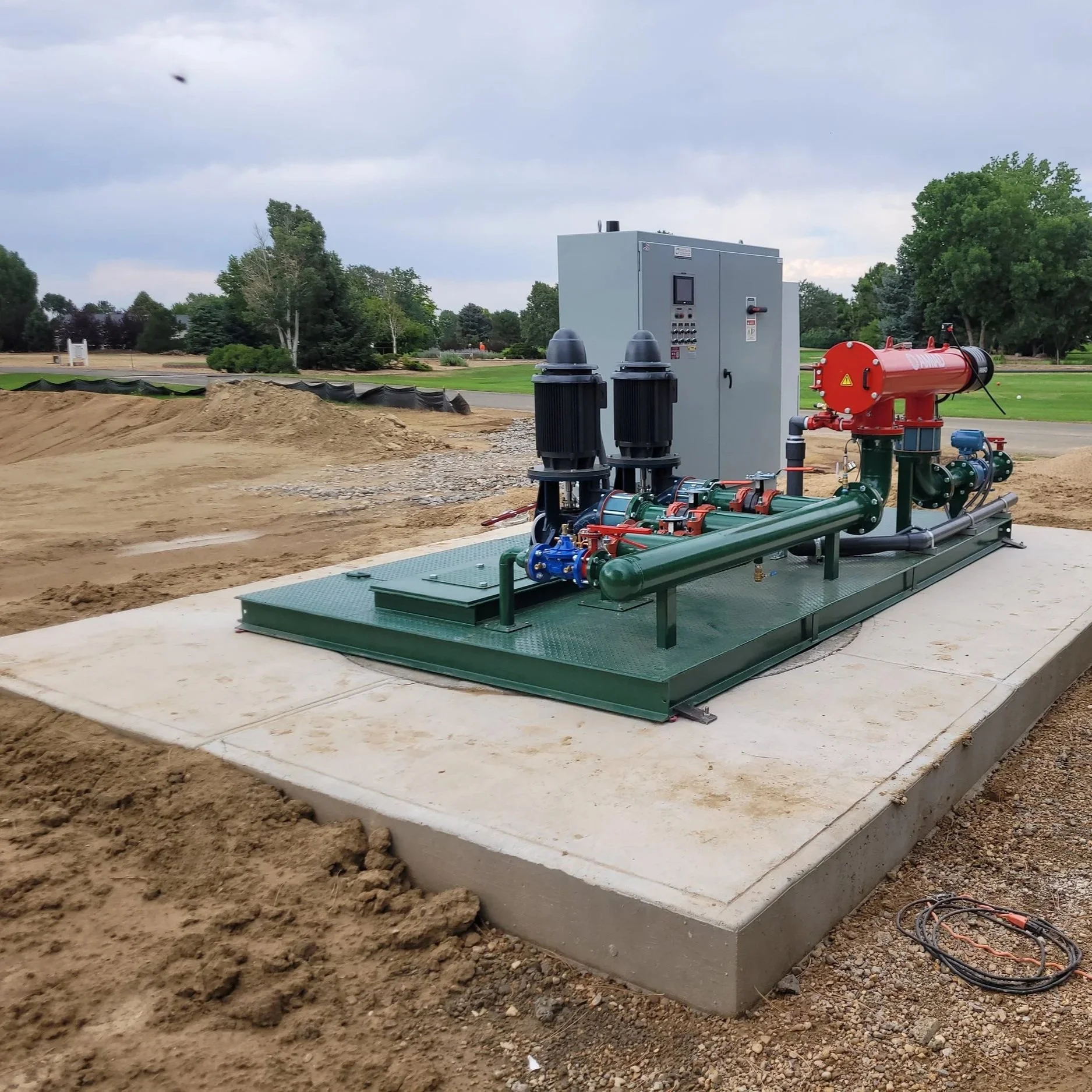 Underground utility equipment on a construction site, including pipes, a control panel, and a red cylindrical tank, with soil and earthwork in the foreground, trees and a park in the background.