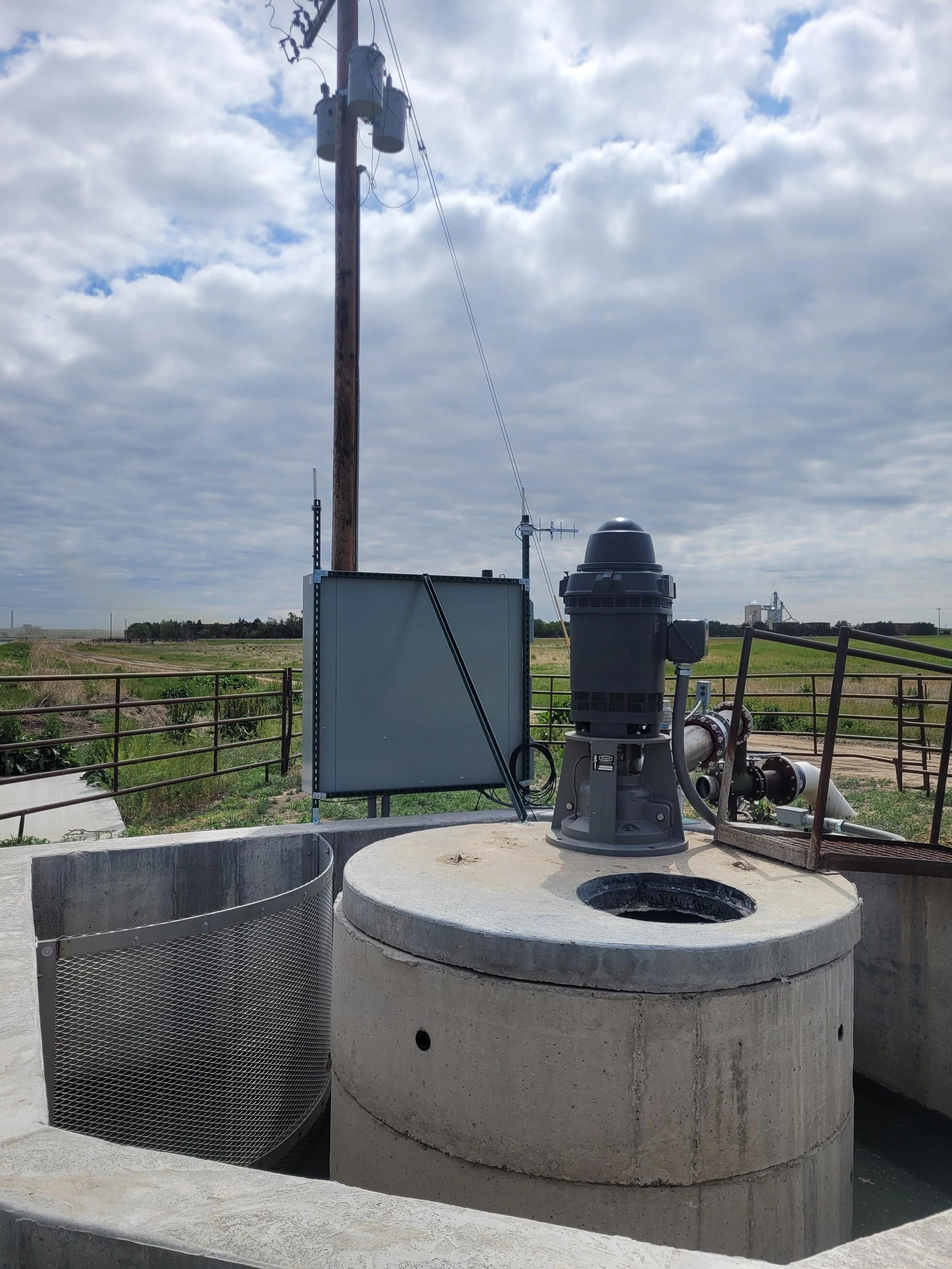 A well pump system outdoors, including a large concrete water well with a motor and pipes, and a utility box, against a background of a cloudy sky and open fields.