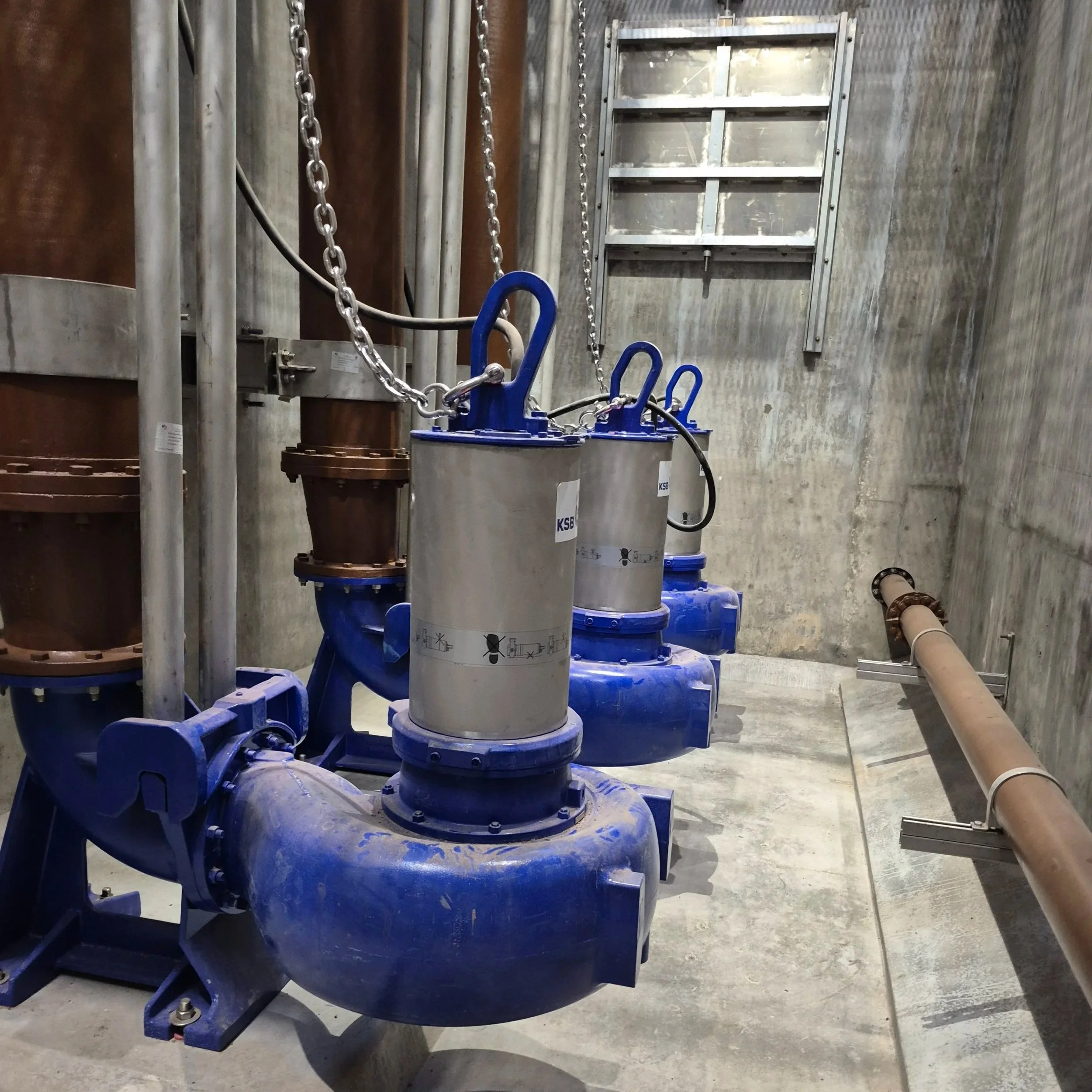 Large blue industrial pumps connected to pipes and chains in a concrete room, possibly part of a water or wastewater treatment system.
