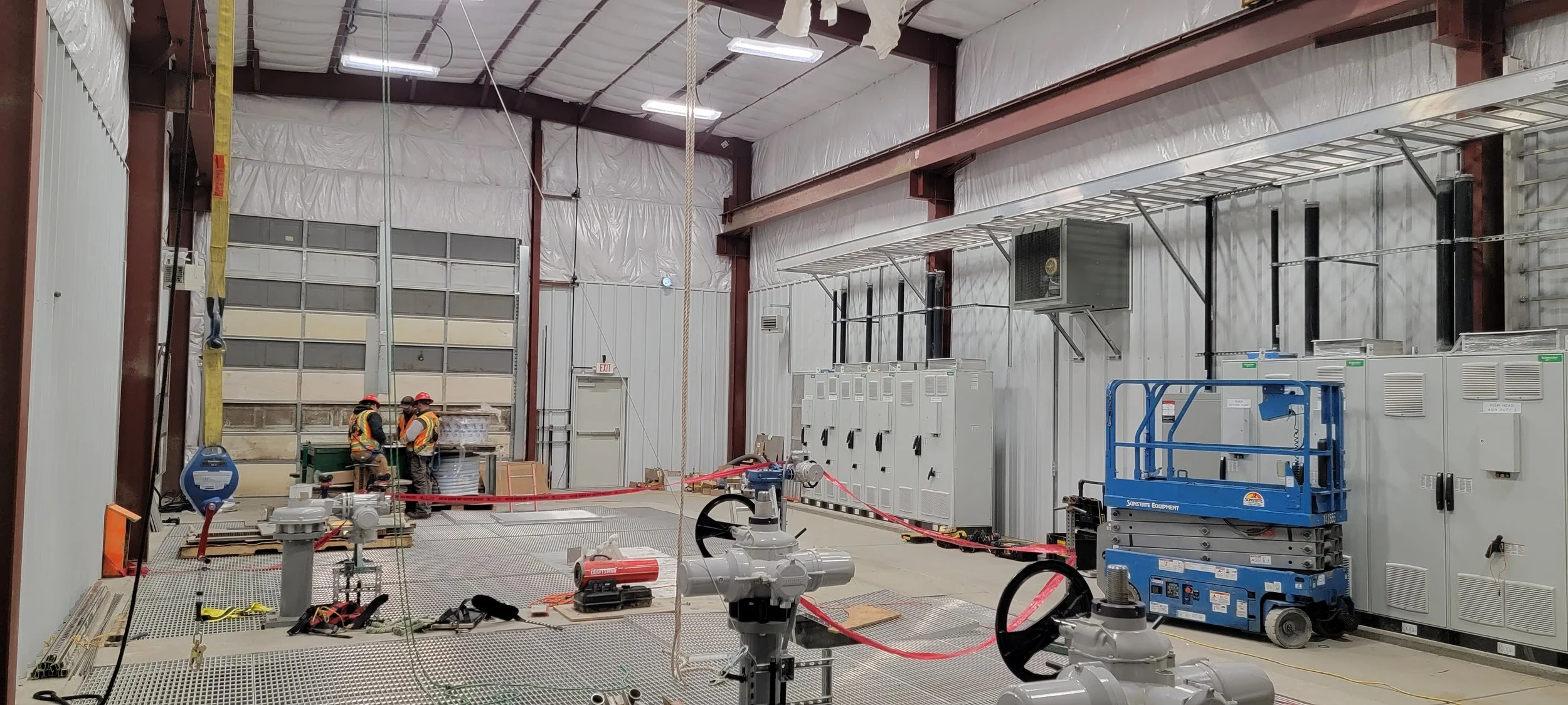 Three construction workers wearing helmets and safety vests working inside a warehouse or industrial facility with various equipment, tools, and machinery around.