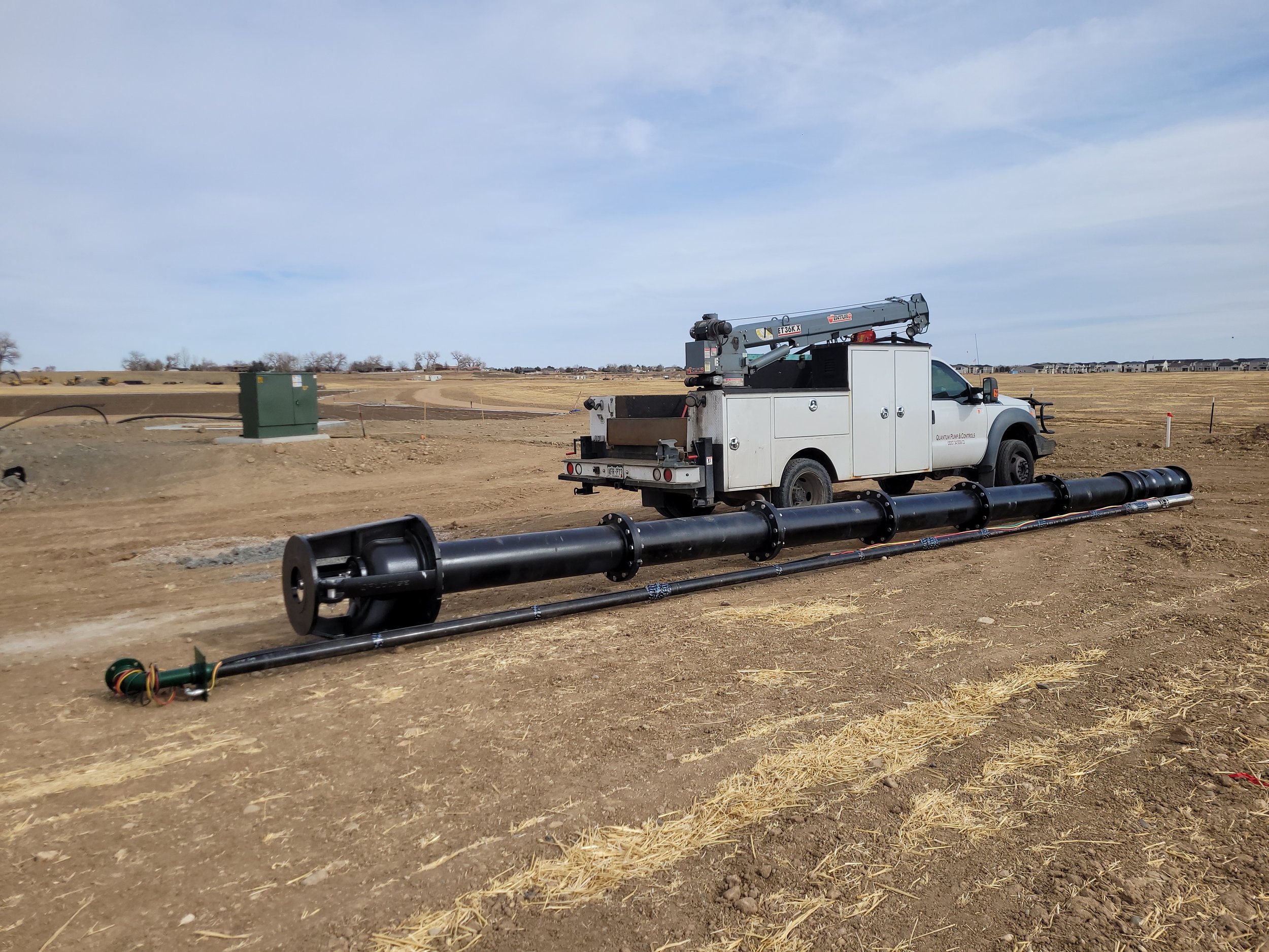 Construction site with a white utility truck equipped with a crane, and large black pipes on the ground, under a partly cloudy sky.