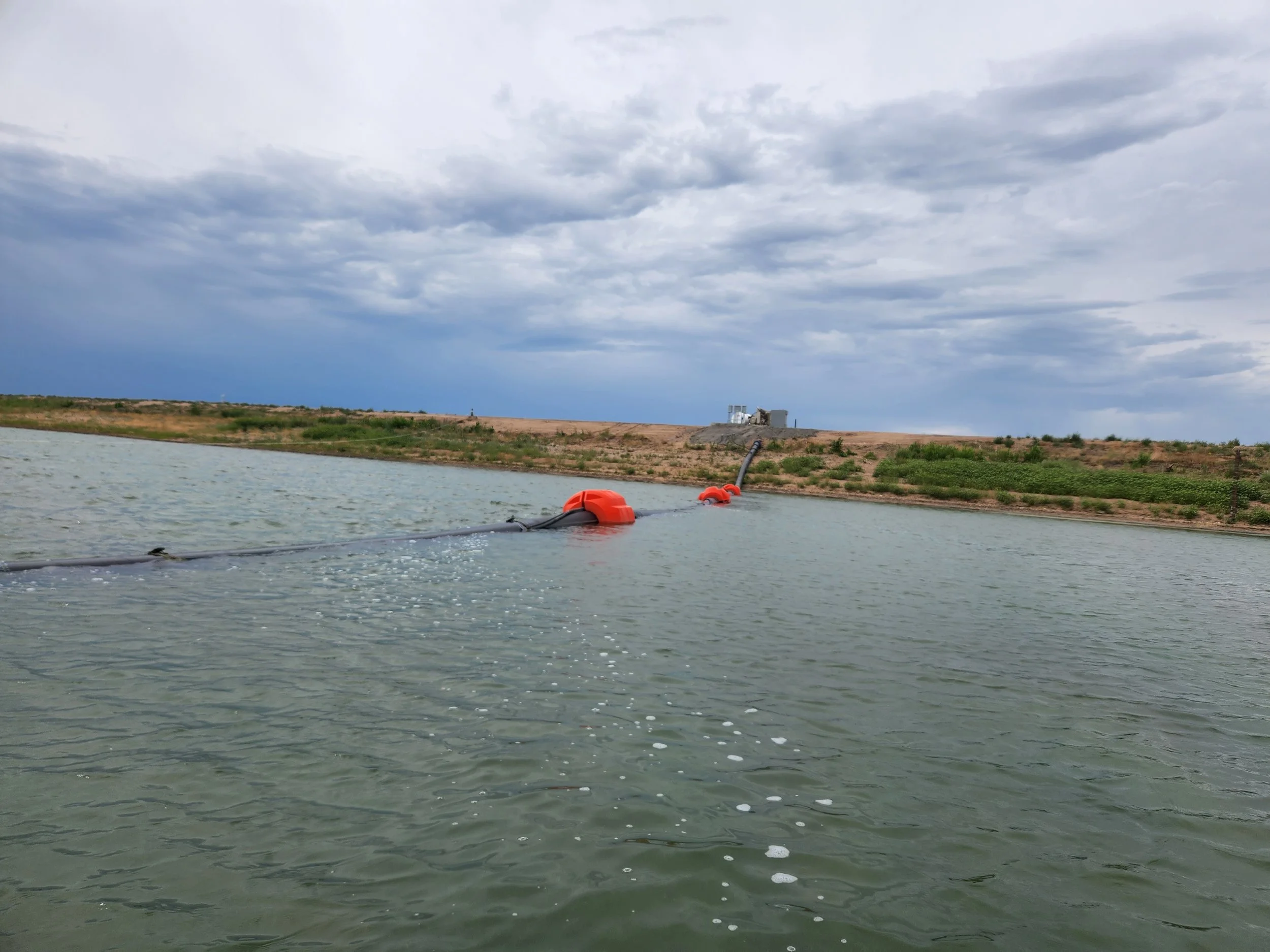 A body of water with a floating pipeline and a distant shoreline with some structures and cloudy sky.