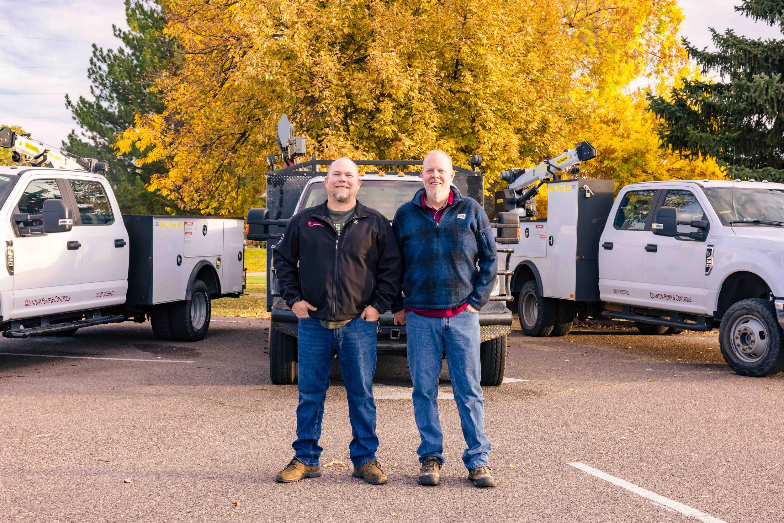 Two men standing in a parking lot in front of three utility trucks, with large trees with yellow leaves in the background, indicating fall season.