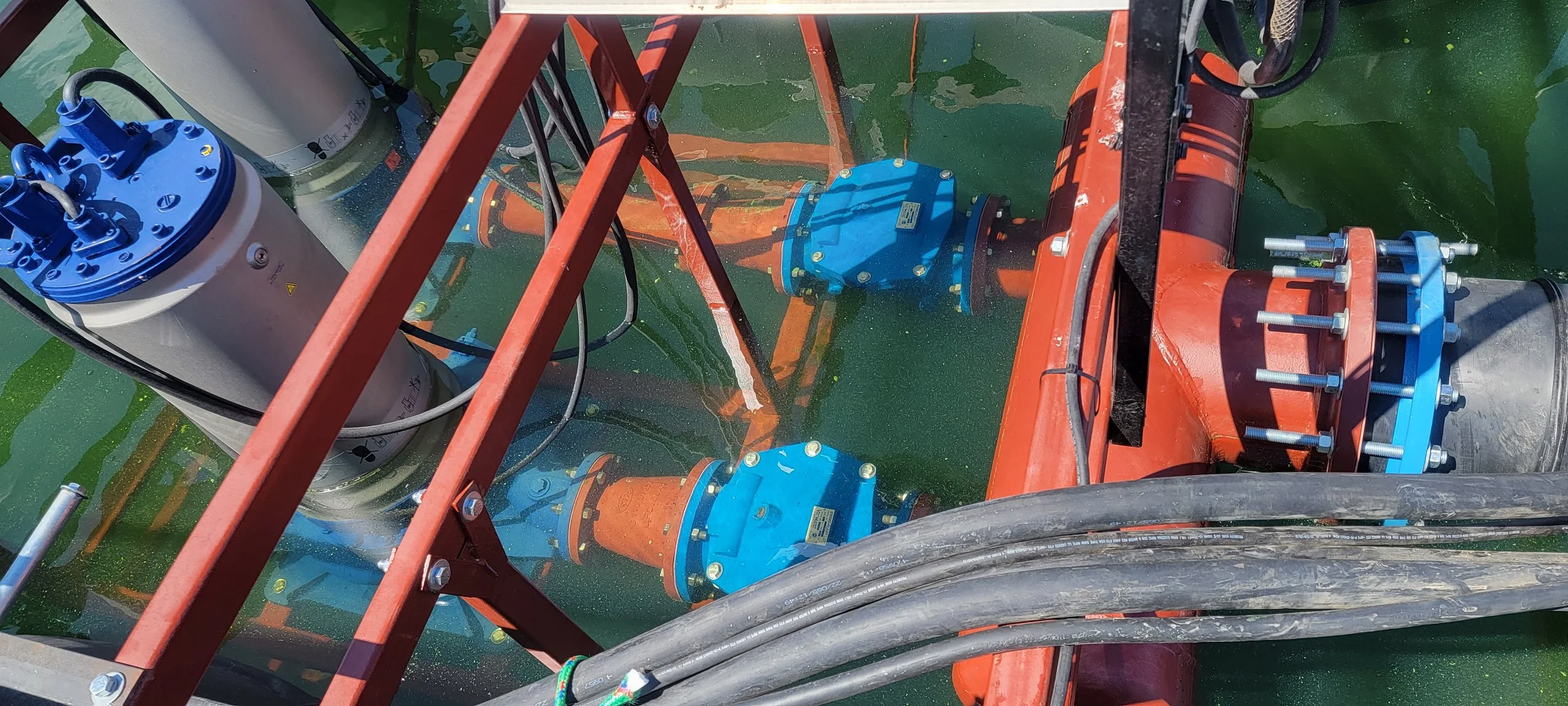 Close-up view of underwater industrial equipment with orange and blue painted metal parts, bolts, pipes, and hydraulic or pneumatic connections submerged in green water.