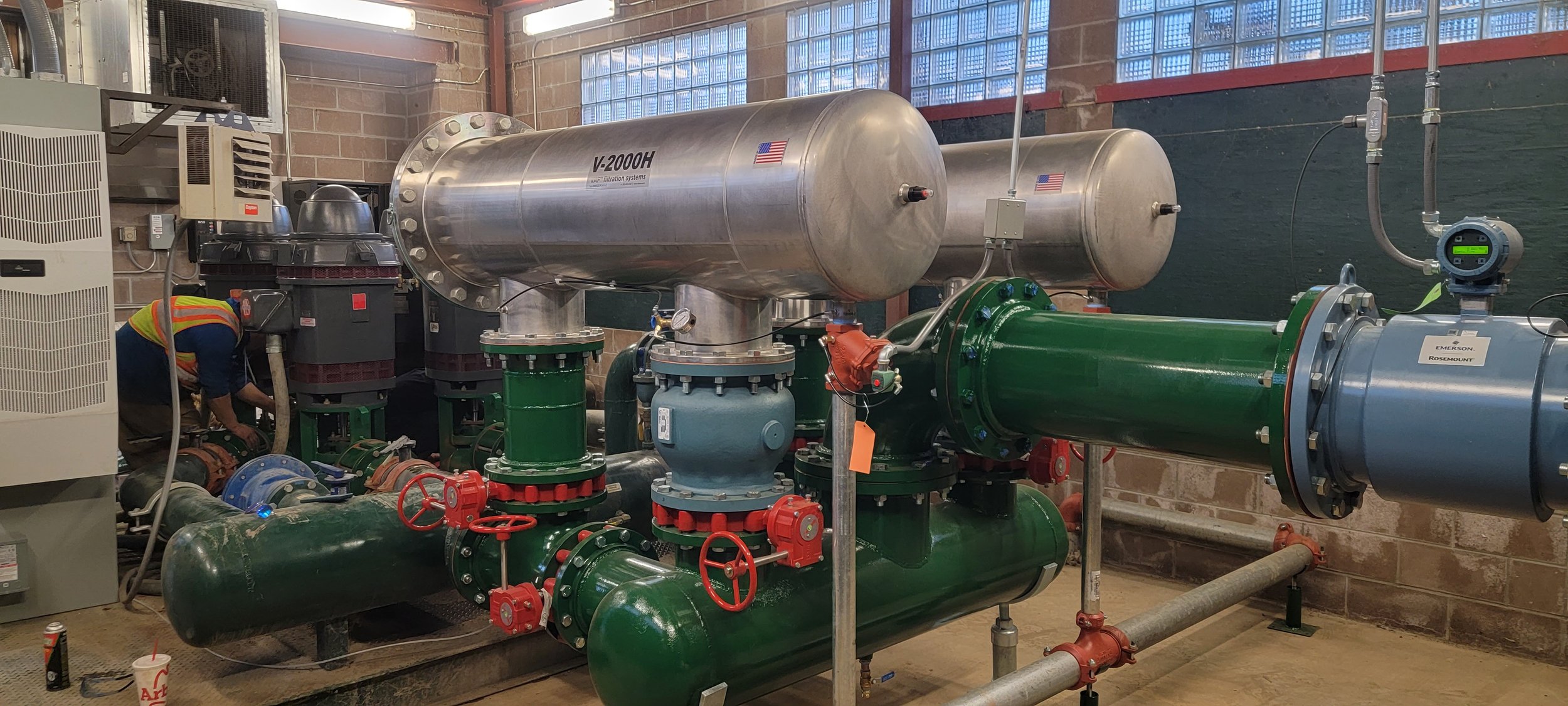 An industrial room with large green and silver pipes, valves, and machinery used for water or fluid systems. A worker is adjusting equipment on the left side. The room has brick walls and glass block windows.