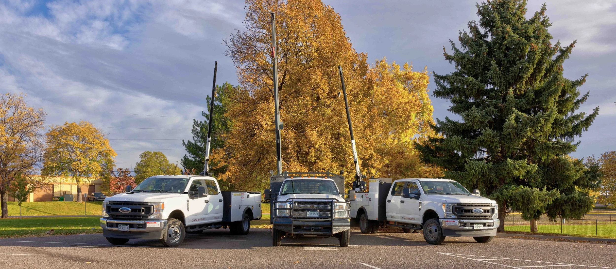 Three utility trucks with buckets raised, parked in a parking lot on a fall day with trees showing autumn colors in the background.