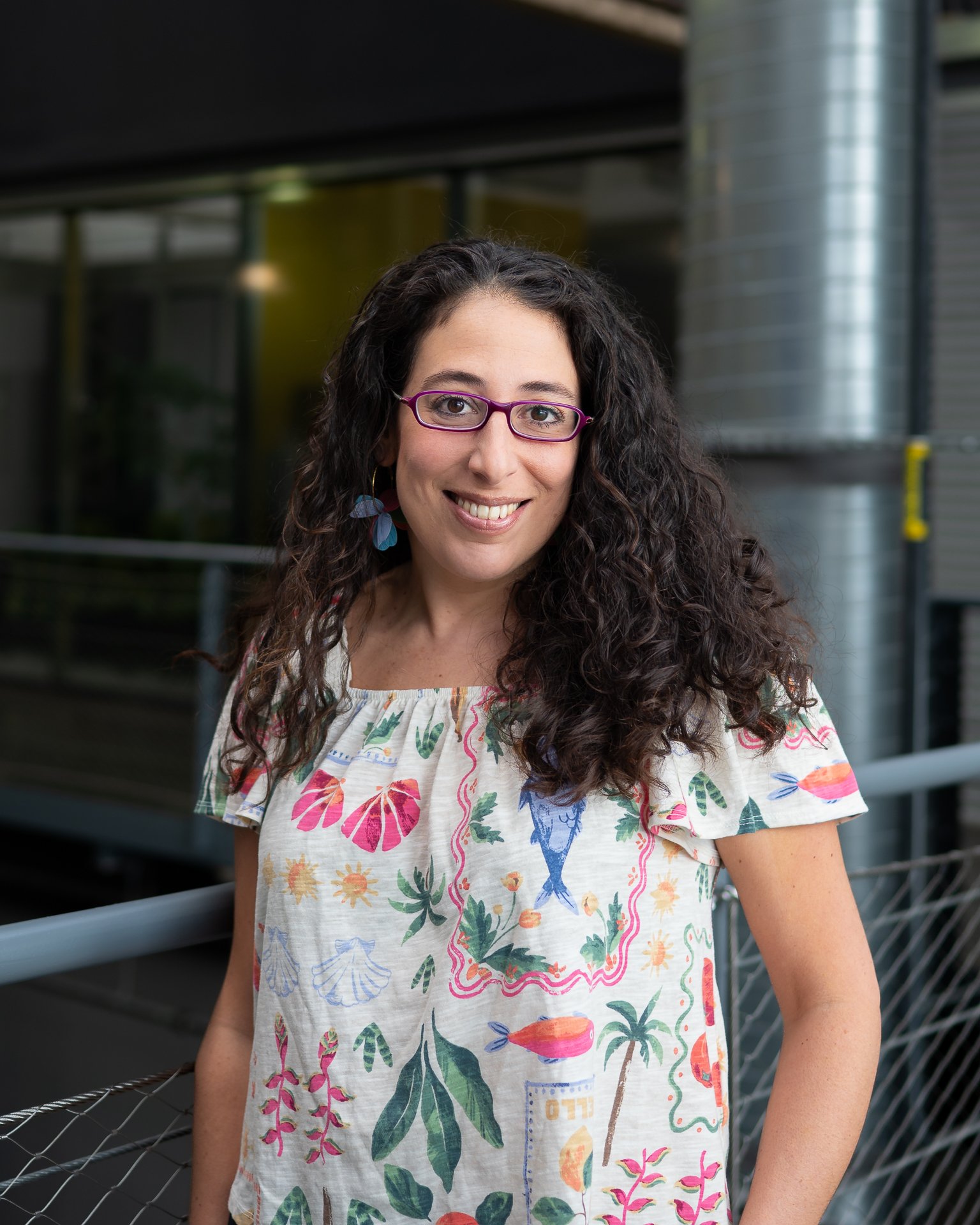 Une femme souriante portant des lunettes violettes, avec des cheveux bouclés foncés, portant une robe à motifs floraux et colorés, posant à côté d'un garde-corps en métal dans un environnement urbain moderne.