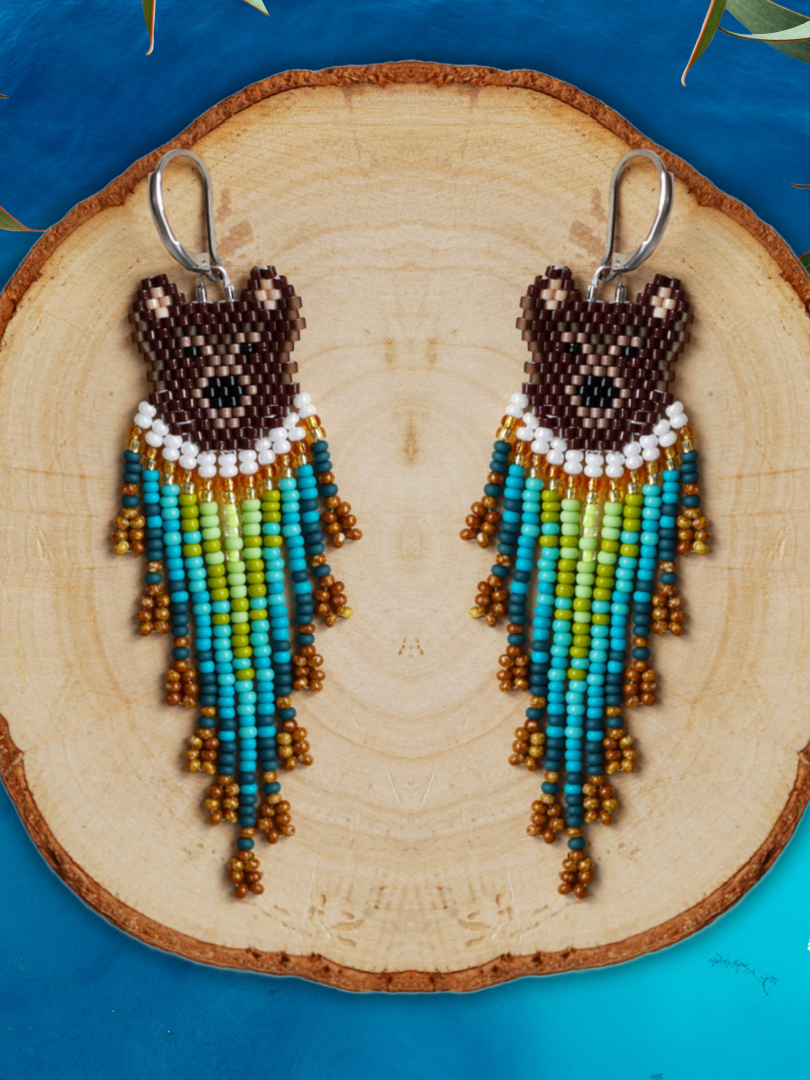Boucles d'oreilles en perles représentant un visage de ours avec un collier de perles blanches et une frange de perles colorées, posées sur une rondelle de bois avec un fond bleu.