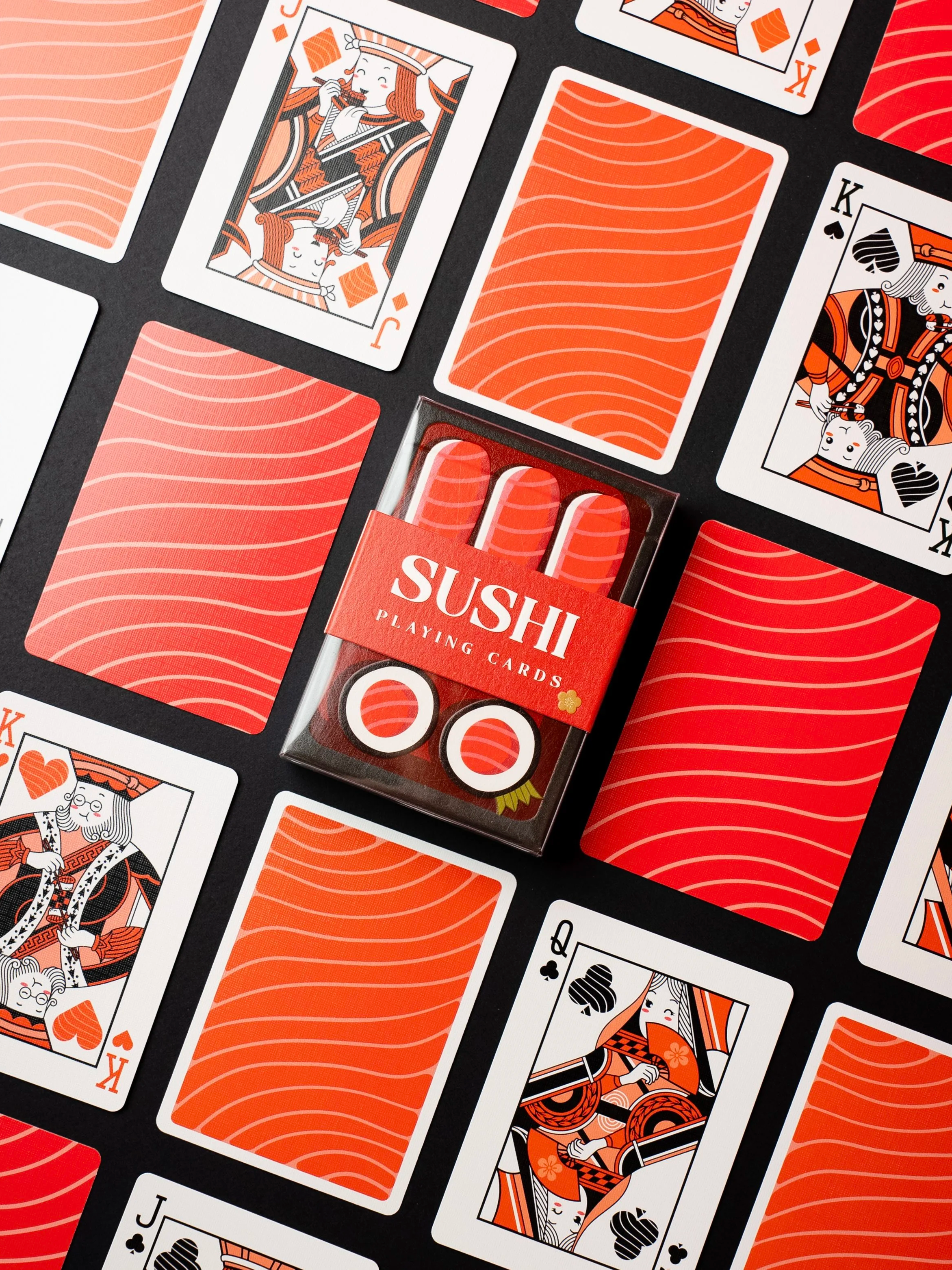 Jeu de cartes de sushi avec cartes colorées en rouge, blanc et noir, représentant des dessins de style japonais, et un paquet de cartes nommé 'SUSHI PLAYING CARDS'.