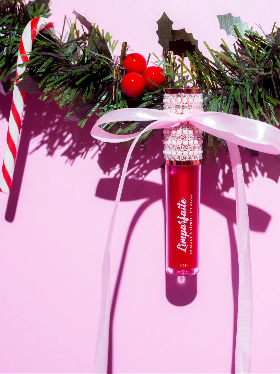 Baume à lèvres rouge nommé 'Cinnamon', avec un ruban rose, accroché à une décoration de Noël composée de sapin en plastique, d'une branche de houx avec des berries rouges, et une sucette de Noël à rayures rouges et blanches, sur un fond rose.