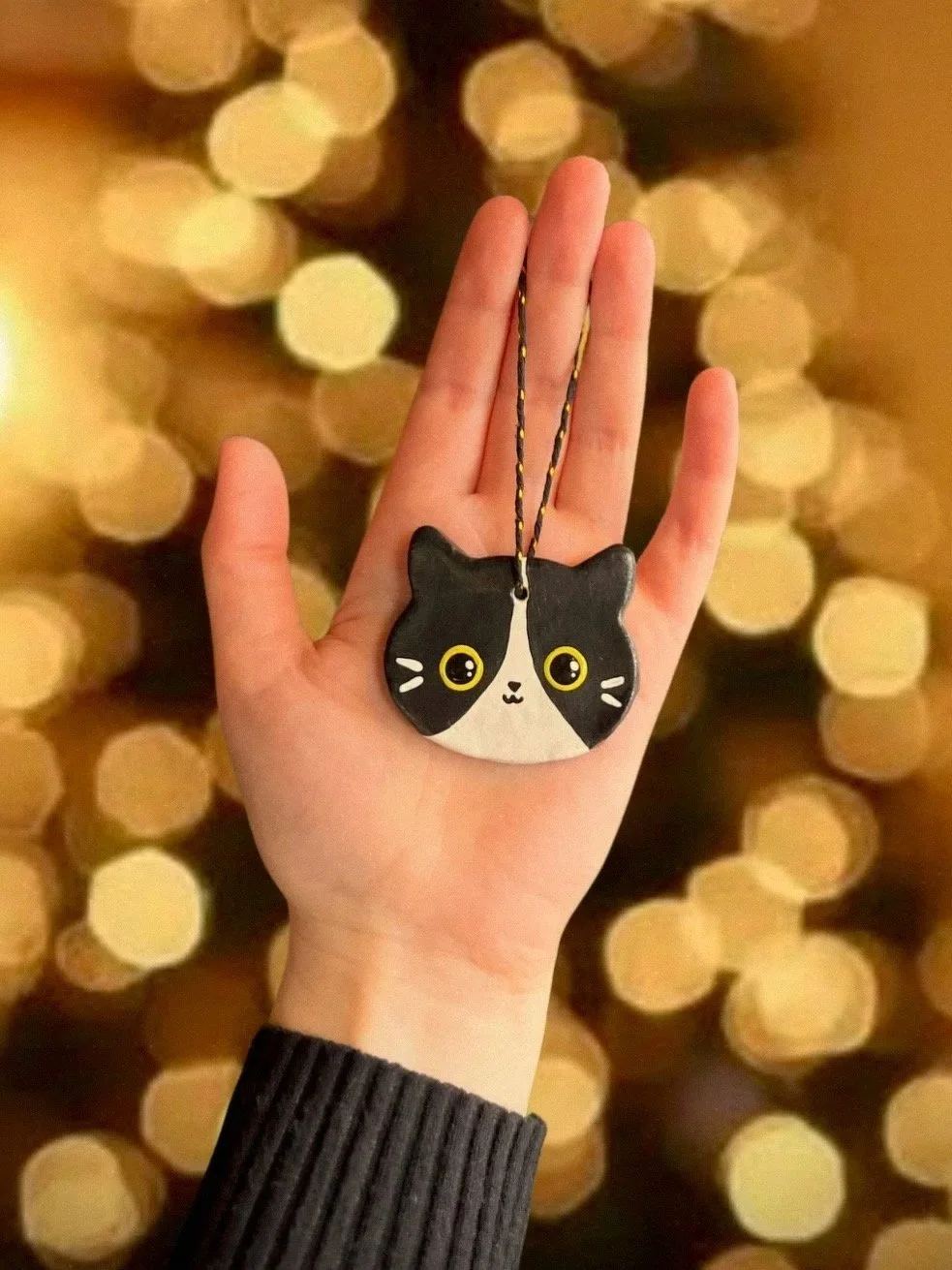 Main tenant une main portant un porte-clés en forme de chat noir et blanc avec de grands yeux jaunes, un fond flou de lumières dorées