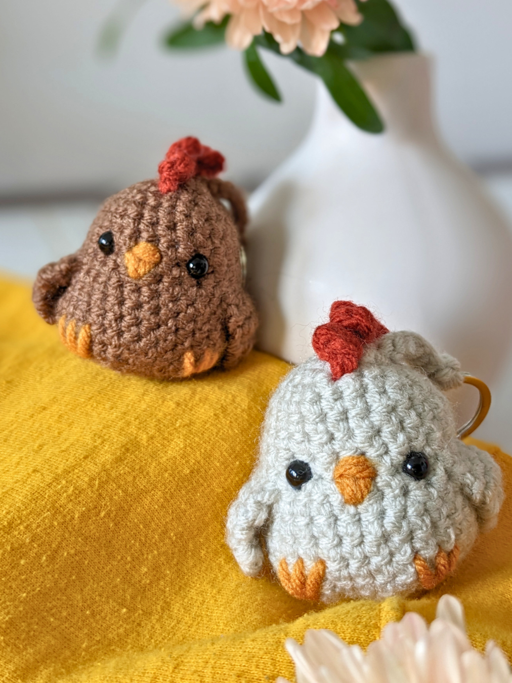 Deux poules en peluche crochetées, une brune et une blanche, posées sur un coussin jaune, avec une vase blanche contenant des fleurs en arrière-plan.
