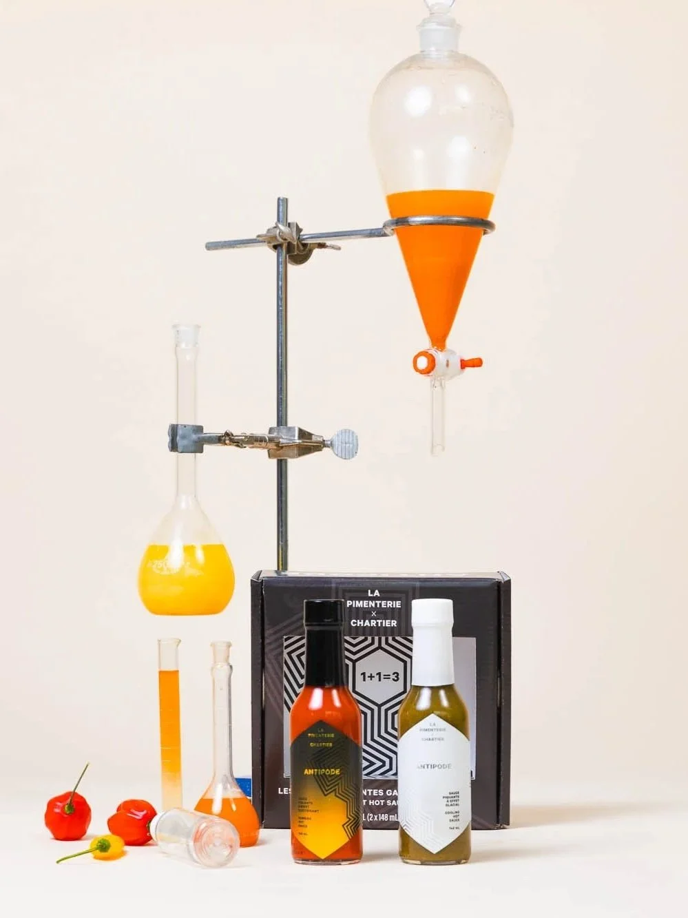 Matériel de laboratoire avec bouteilles contenant des liquides orange et jaune, fioles, petits piments rouges, boîte noire avec texte, et éléments de chimie