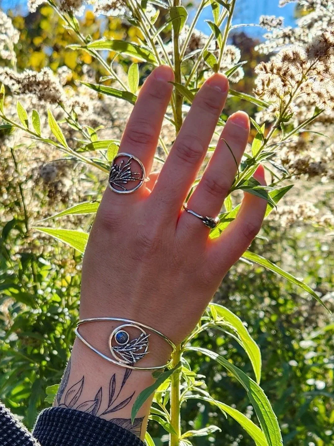 Une main portant plusieurs bagues en argent avec des motifs de feuilles, tenant une branche verte avec de petites fleurs blanches, en arrière-plan un ciel bleu clair et d'autres plantes.