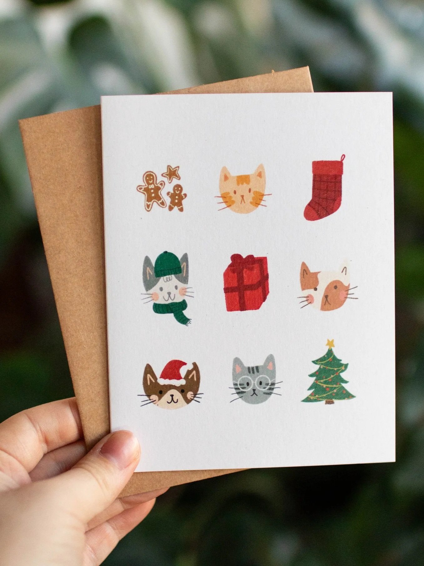 Une carte de vœux de Noël avec des dessins de biscuits, chats, cadeaux, chaussette de Noël, arbre de Noël et autres décorations festives, tenue par une main avec un fond flou de verdure.