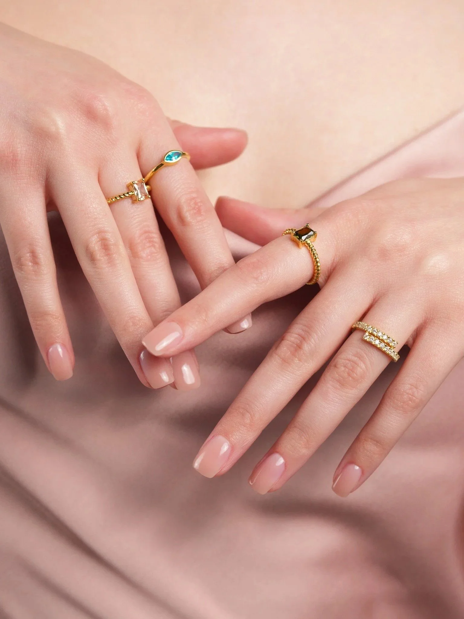 Zoom sur des mains féminines portant plusieurs bagues en or avec des pierres précieuses ou semi-précieuses sur les doigts, posé sur un tissu de couleur rose pâle ou beige.