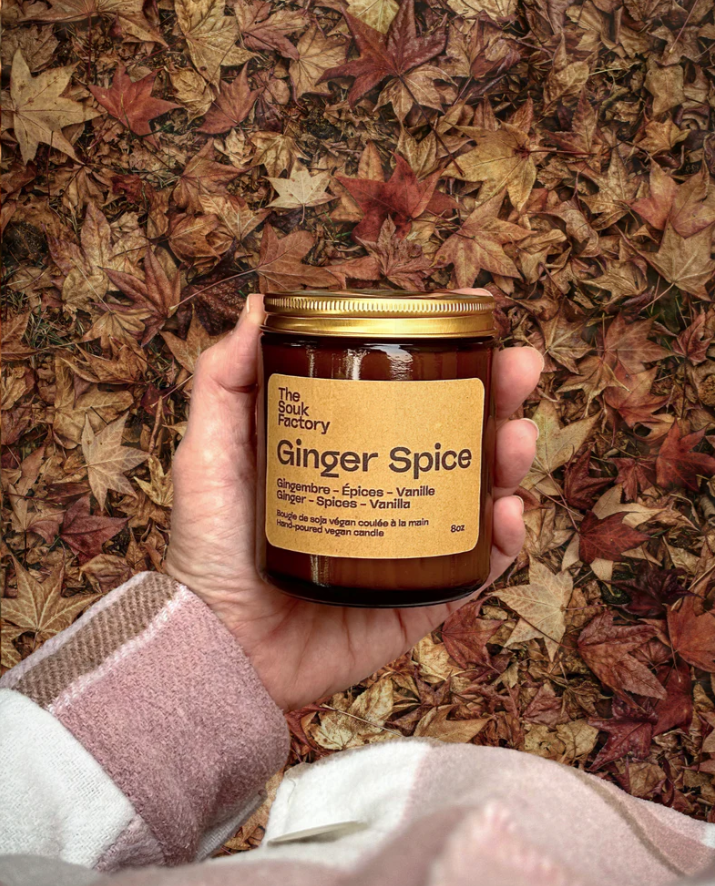 Une main tient un pot de bougie parfumée « Ginger Spice » de la marque The Souk Factory, avec un fond de feuilles mortes d'automne.