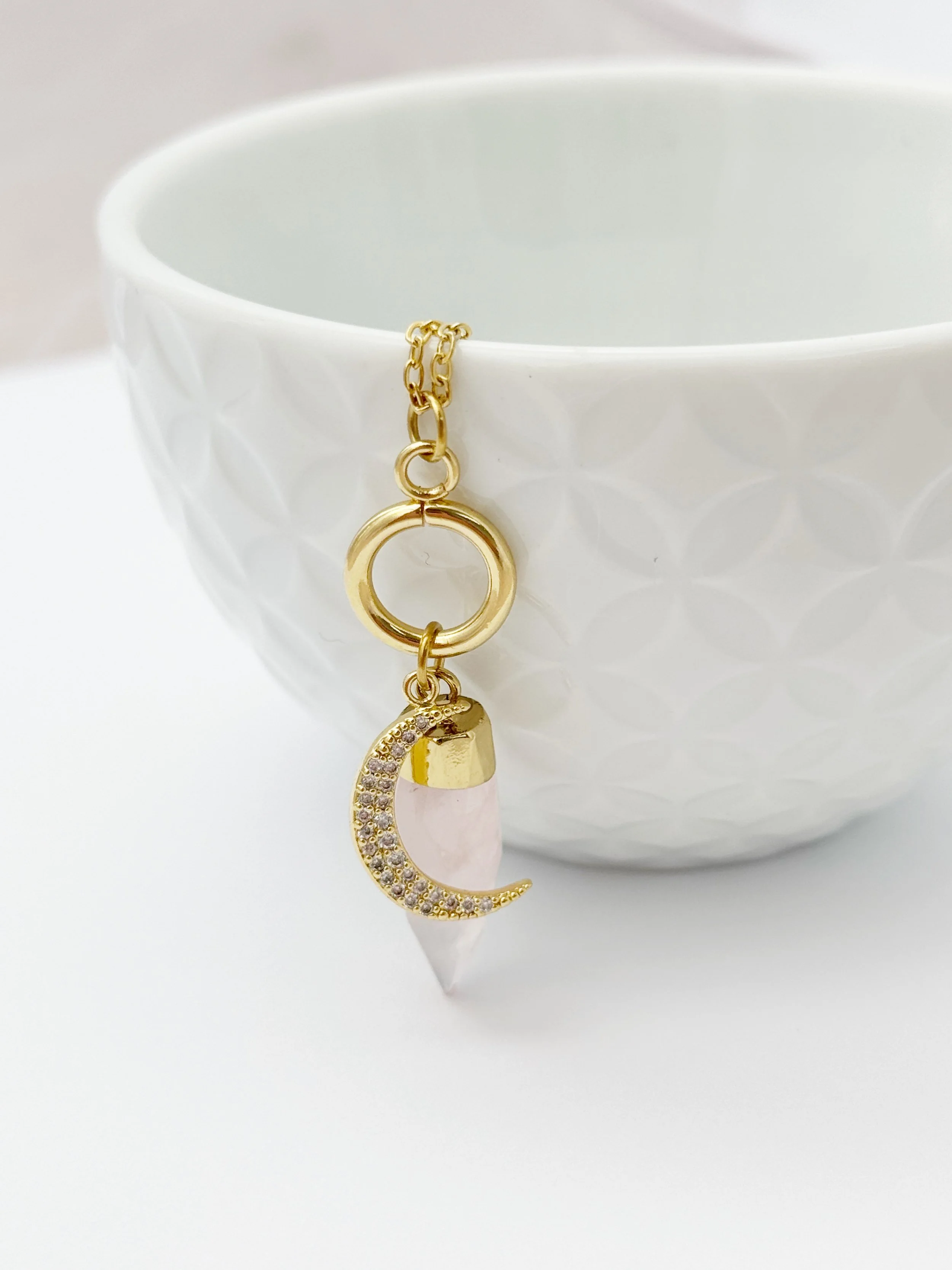 Bracelet orné d'un pendentif en forme de croissant de lune avec des pierres brillantes, suspendu à une chaîne en or, accroché à une coupe blanche