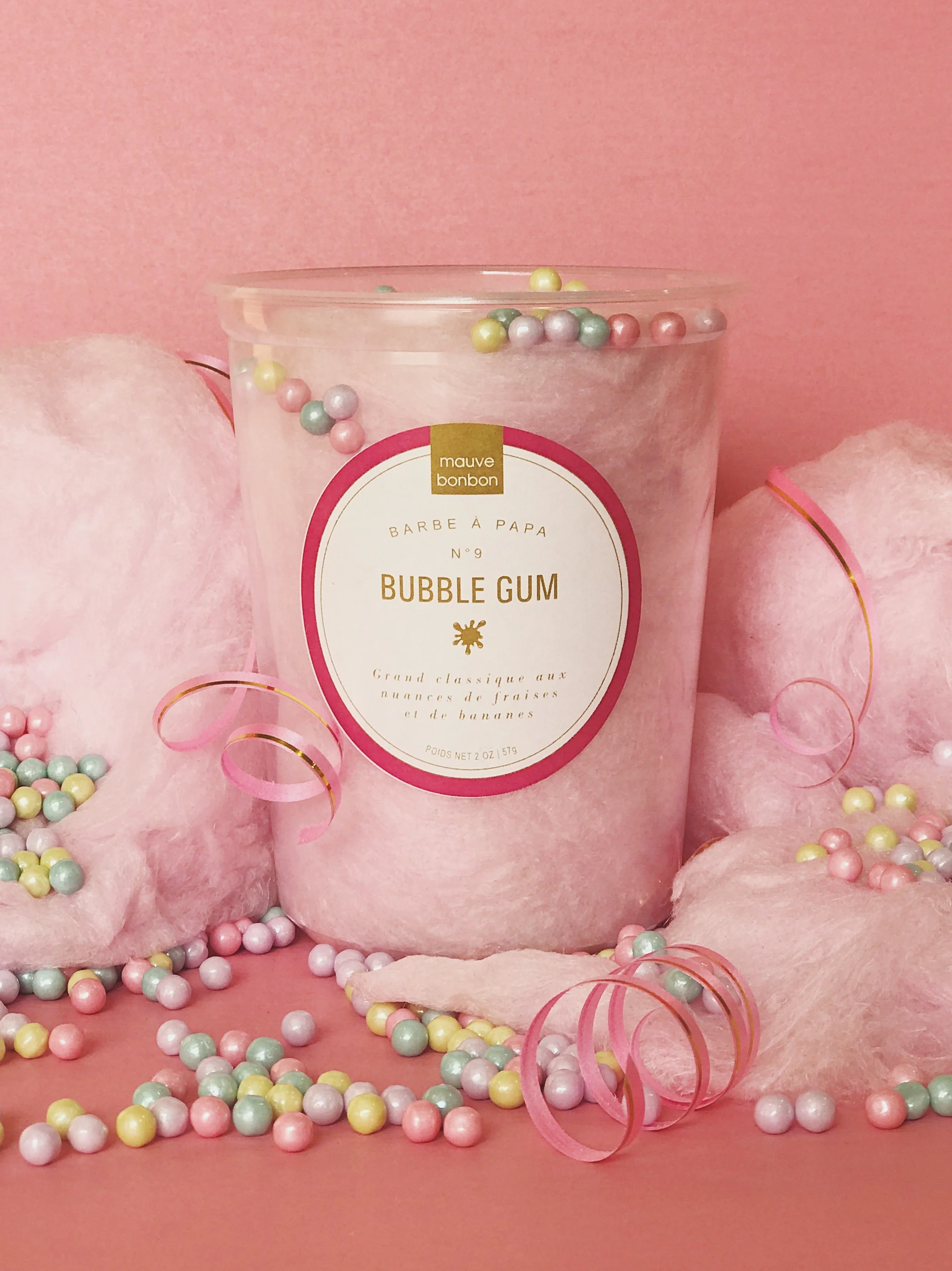 Un pot de crème parfumée aux bonbons de couleur pastel, avec des petites boules en plastique de différentes couleurs autour sur une surface en tissu rose.