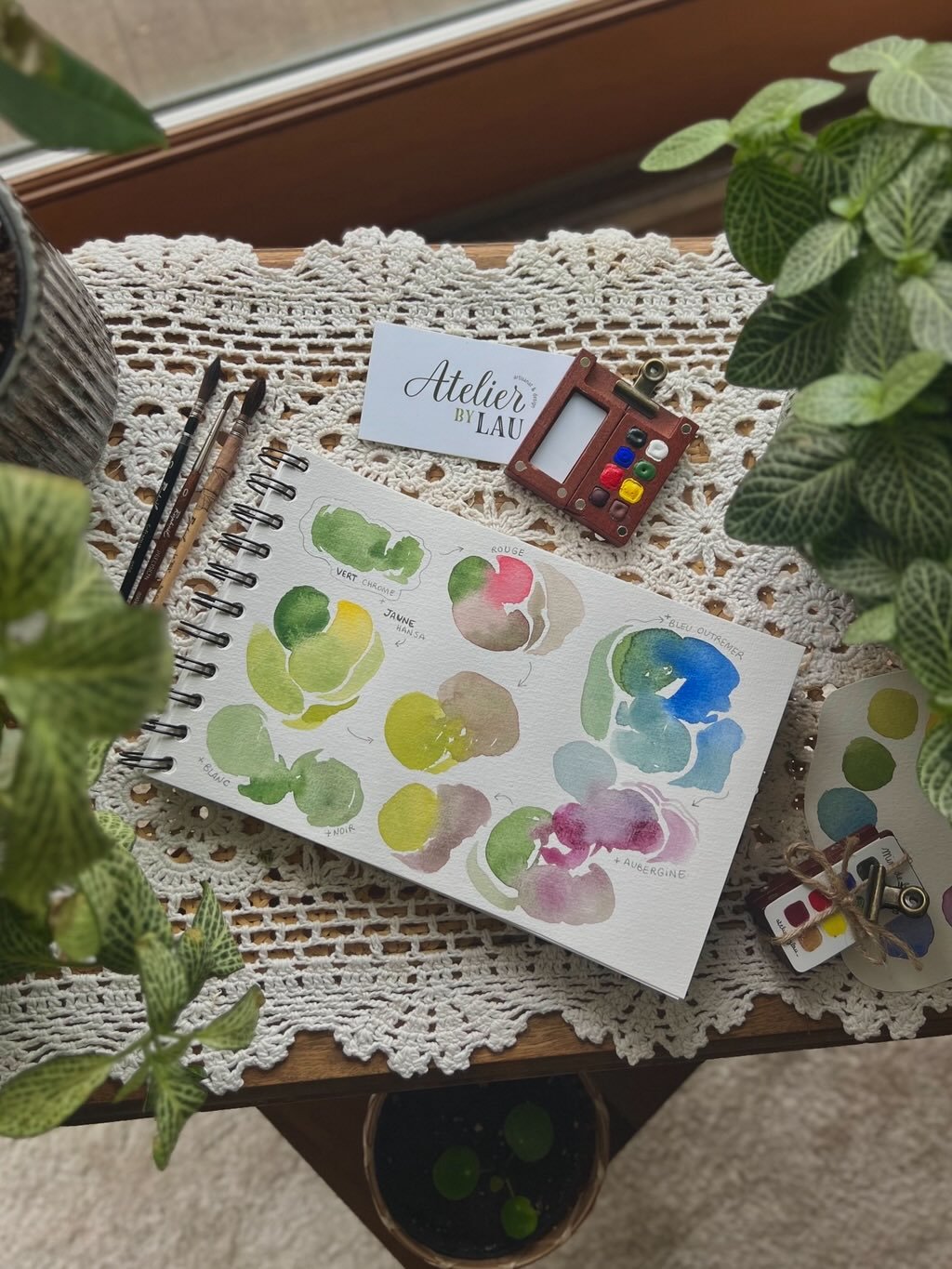 Un carnet de croquis avec des aquarelles de différentes couleurs de fruits, des pinceaux, une petite étiquette "Atelier by Lau", une palette d'aquarelle, et des plantes vertes autour, sur une table recouverte d'une nappe en crochet.