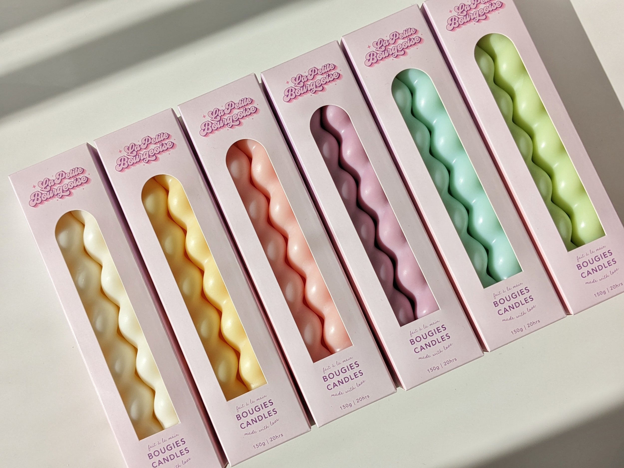 Une rangée de bougies en forme de spirale dans des boîtes roses, de différentes couleurs pastel, avec l'inscription "La Petite Bourgeoise" et "bougies".
